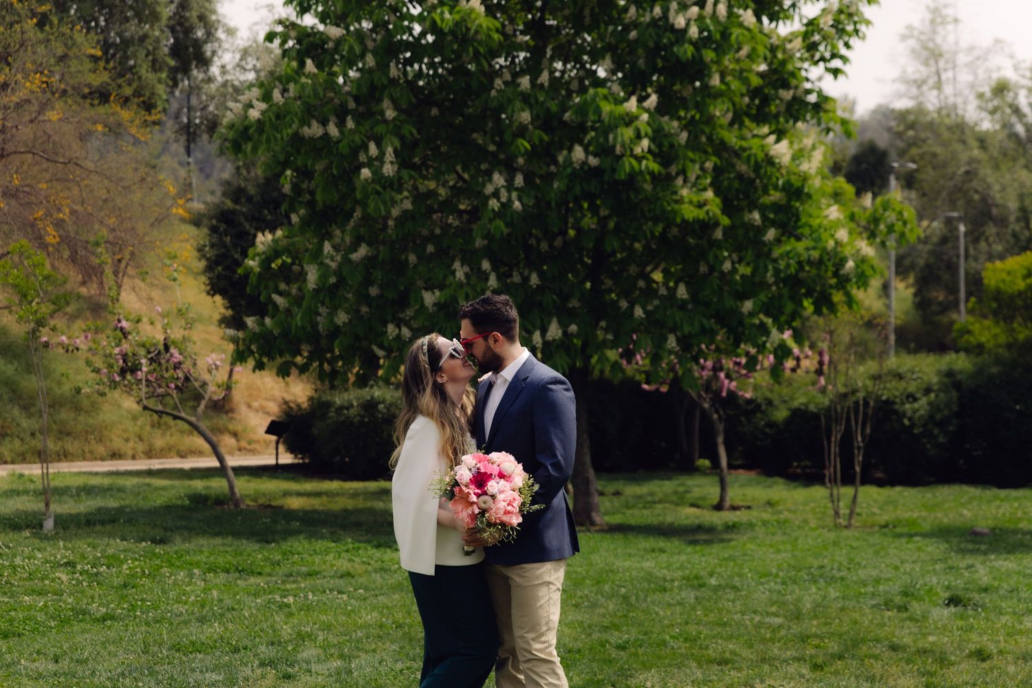 Romantic couple sharing intimate moment on green lawn holding pink bouquet with trees behind.