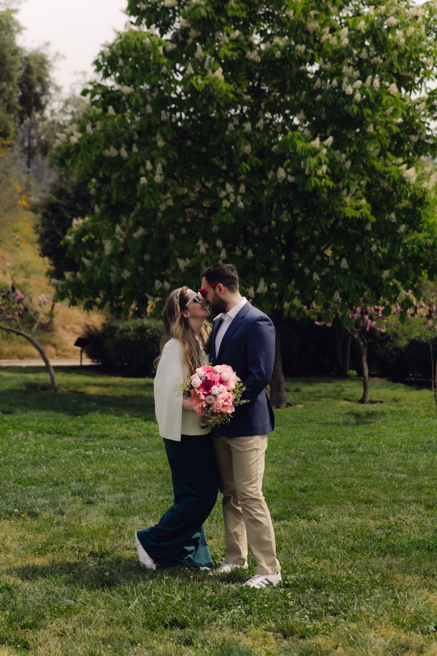 Couple embracing on green lawn with man in blue blazer and woman holding pink bouquet.