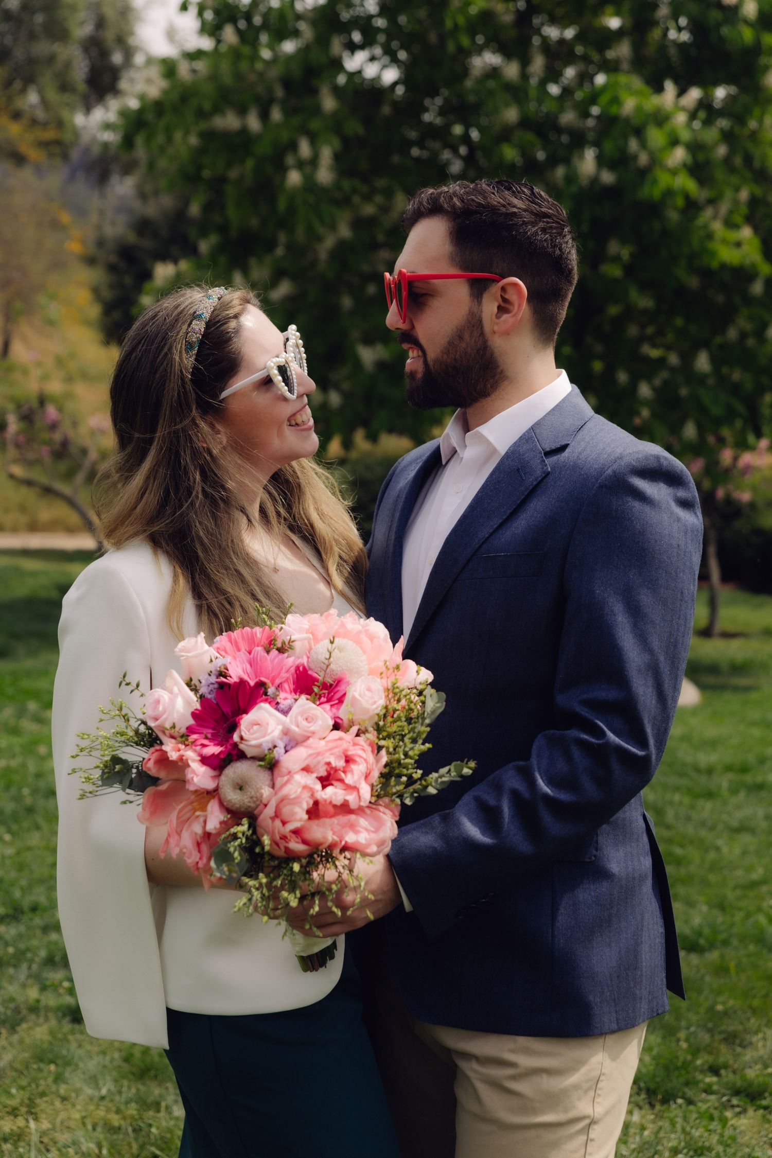 Smiling couple wearing heart-shaped sunglasses holding large pink peony bouquet in green park.