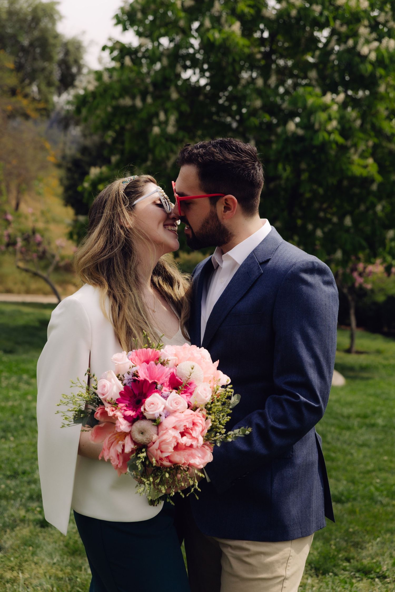 Couple sharing a kiss wearing red heart sunglasses while holding vibrant pink floral bouquet outdoors.