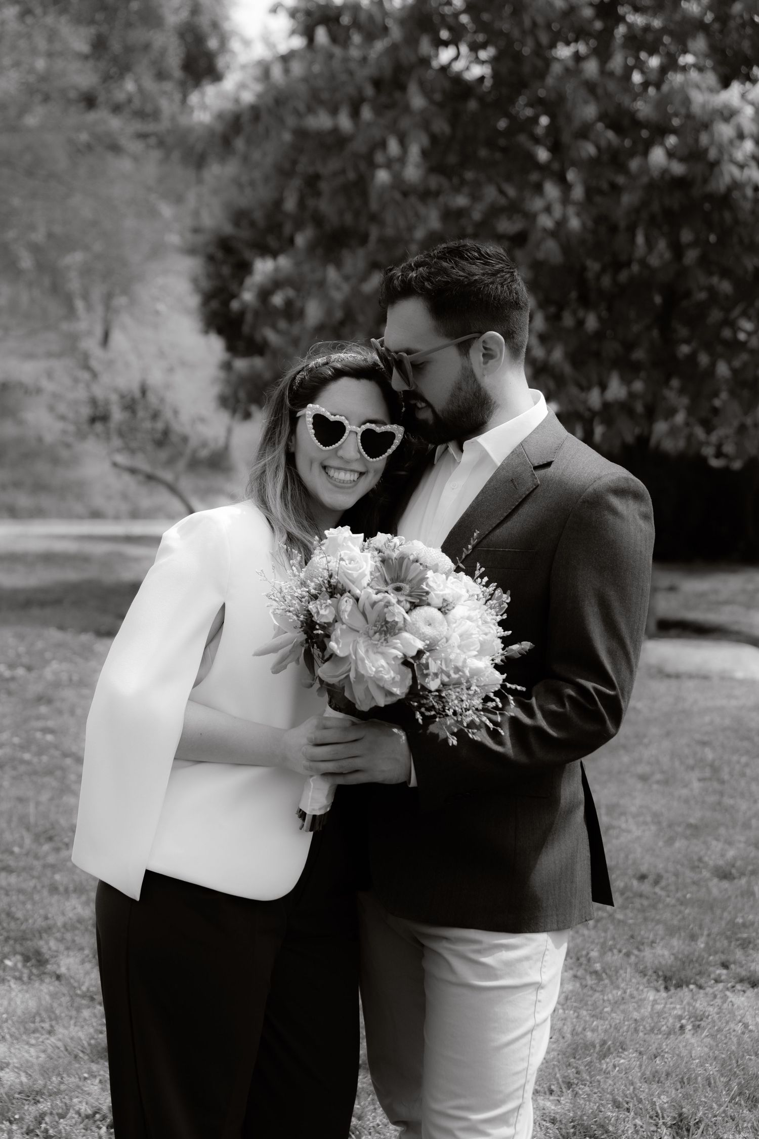 Black and white photo of couple embracing outdoors, woman in white blazer wearing heart sunglasses.