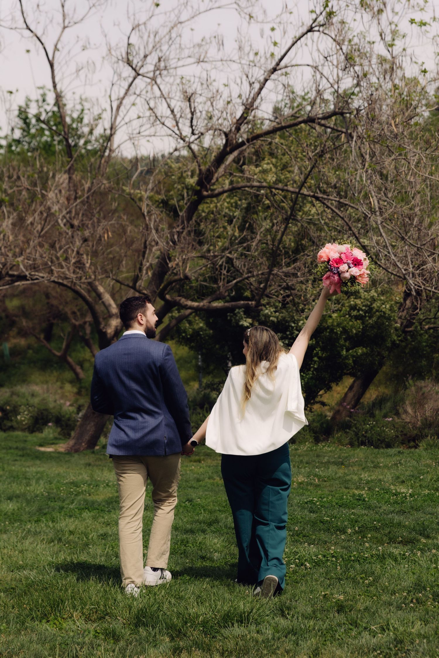 Couple walking hand-in-hand through a green park, bride raising floral bouquet in celebration.