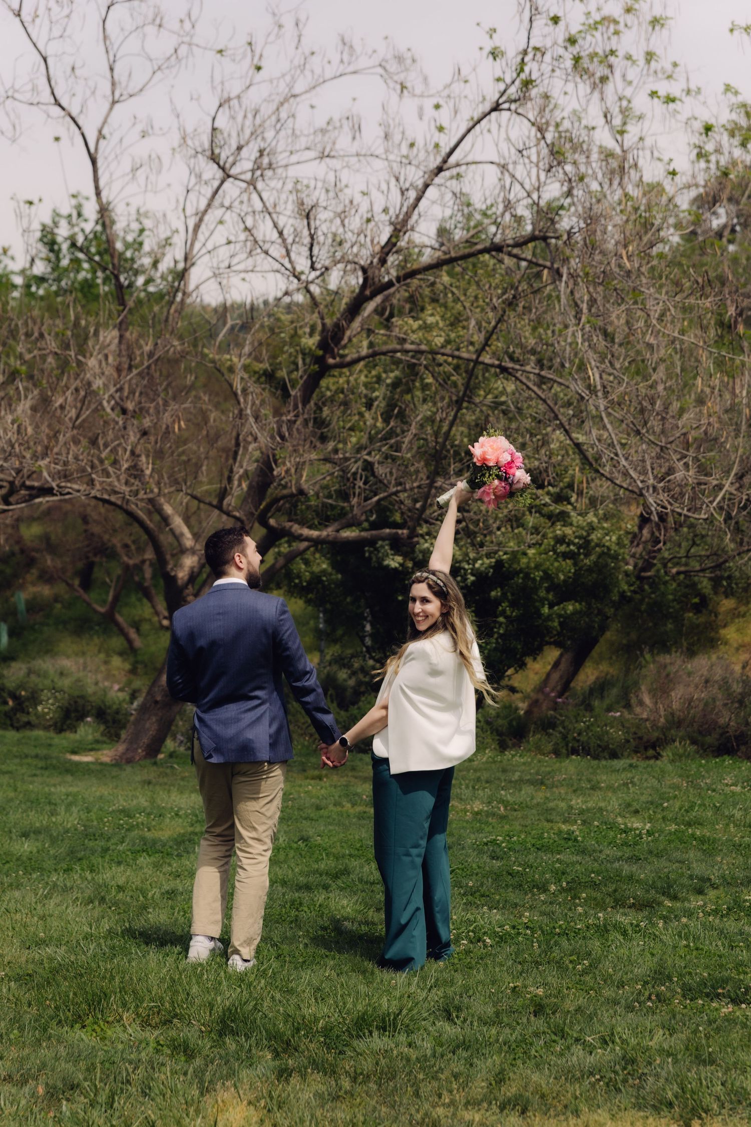 Bride and groom holding hands in a lush garden, woman raising pink bouquet joyfully.