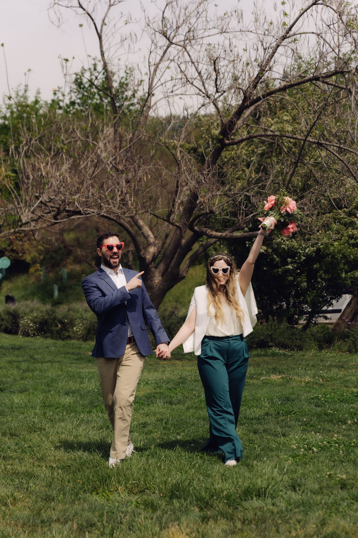 Joyful couple in heart-shaped sunglasses celebrating in a park, bride raising floral bouquet.