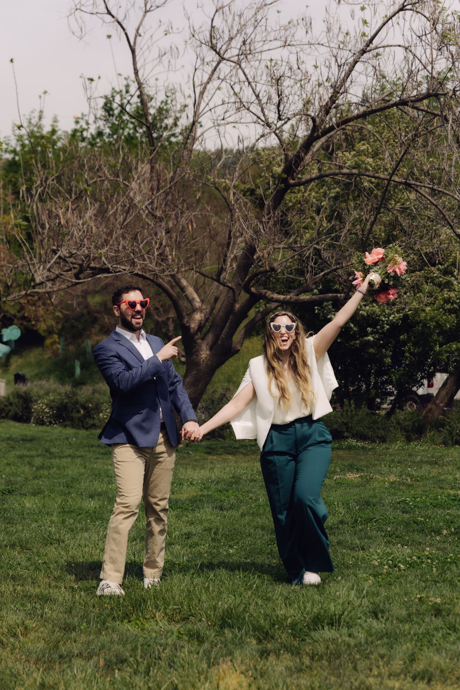 Playful couple wearing fun sunglasses outdoors, bride triumphantly raising pink bouquet in the air.