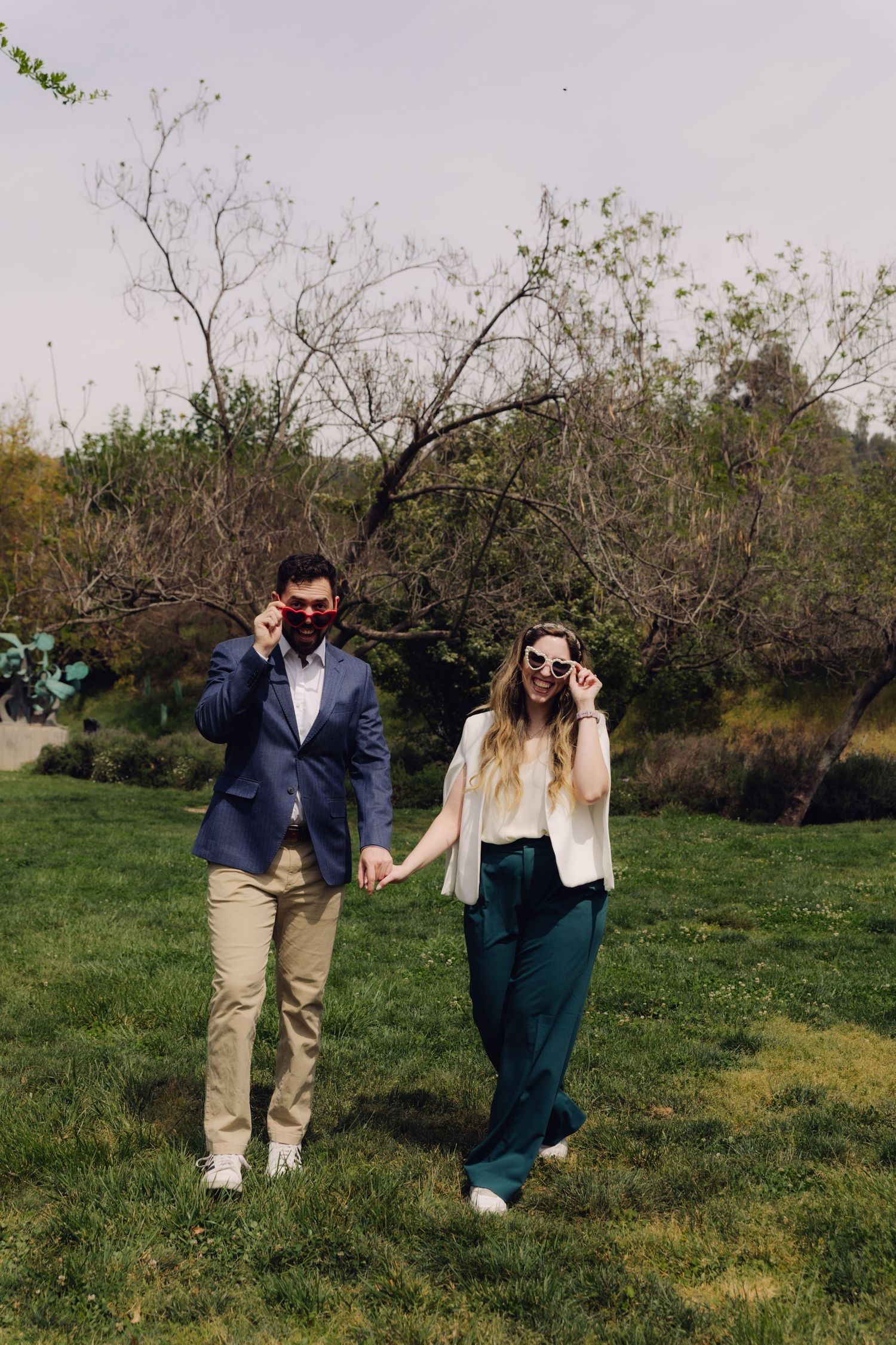 Stylish couple adjusting their sunglasses while walking hand-in-hand through a sunny green park.