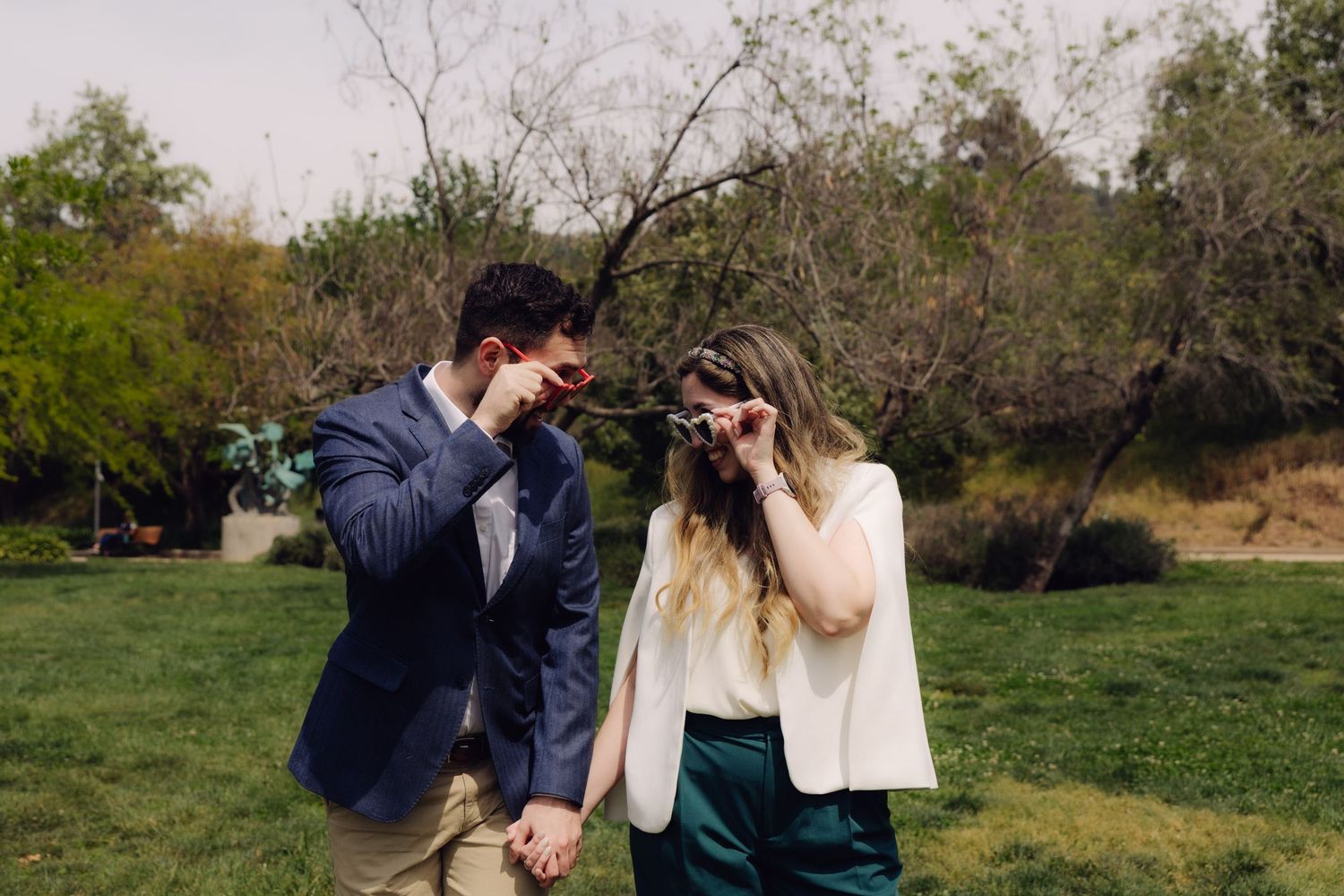 Laughing couple holding hands outdoors, both playfully hiding their faces with sunglasses.