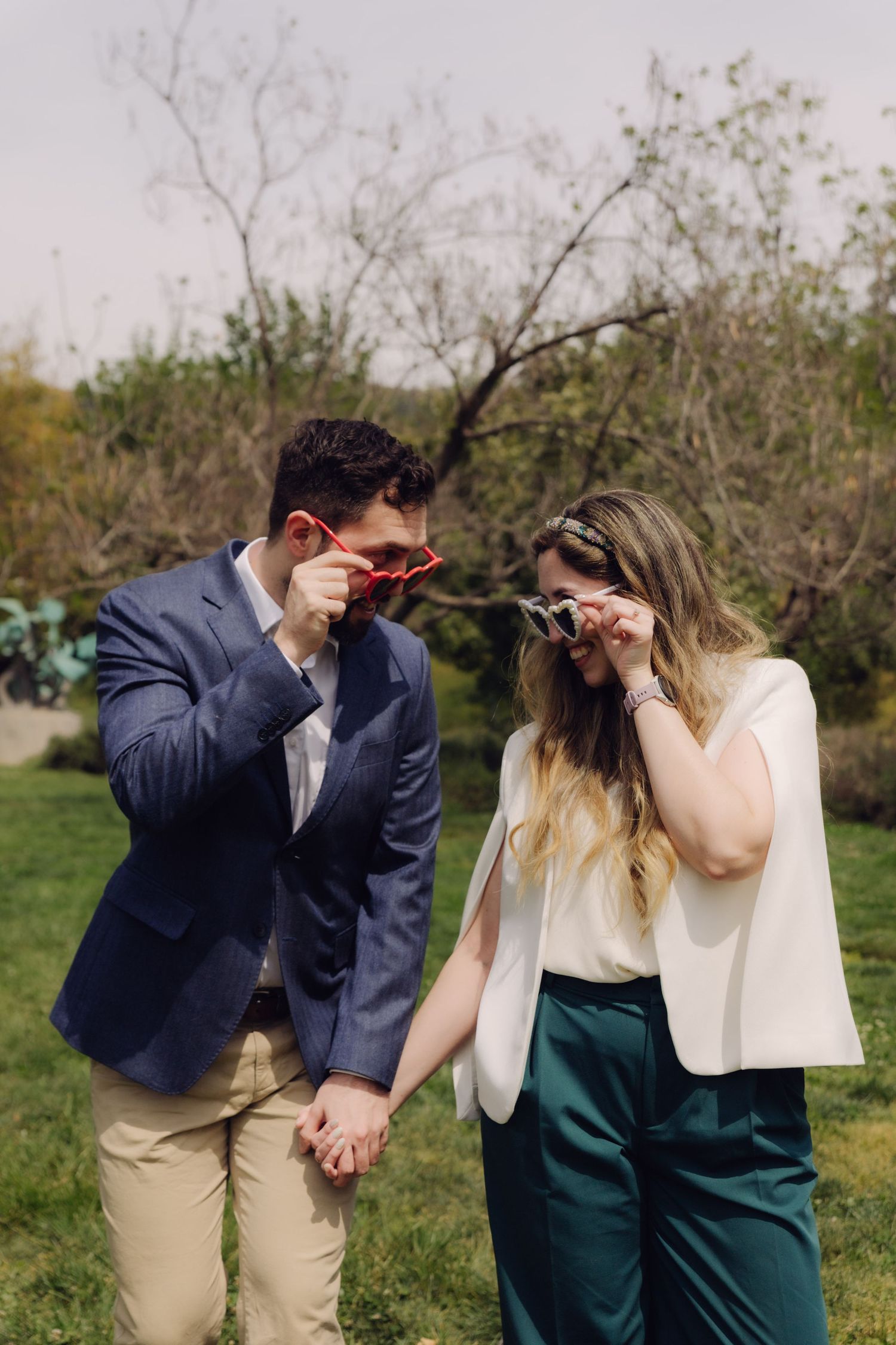 Couple standing in a garden holding hands while adjusting their novelty sunglasses together.