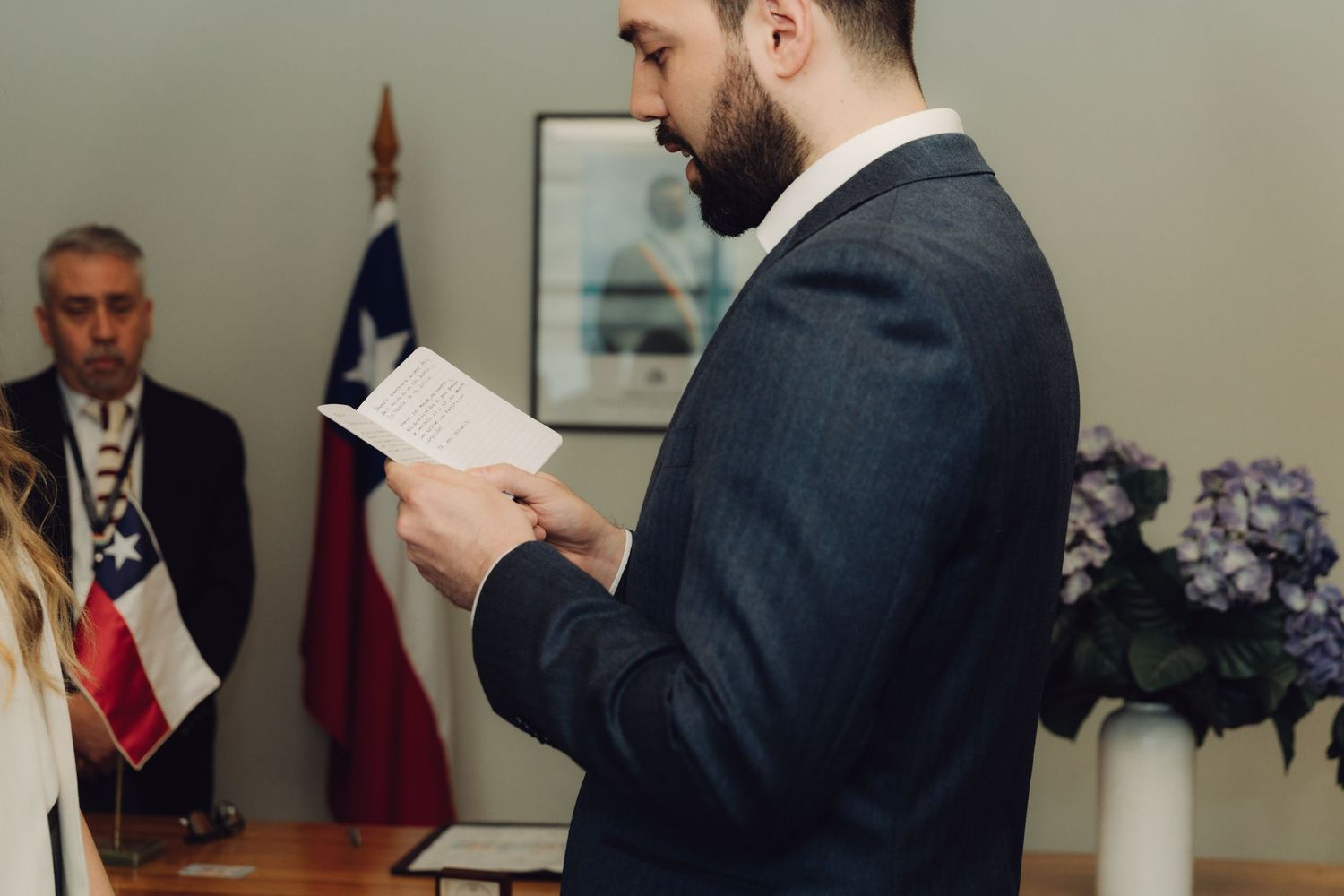 Bearded groom in dark suit reads wedding vows from paper during civil ceremony with Chilean flag in background.