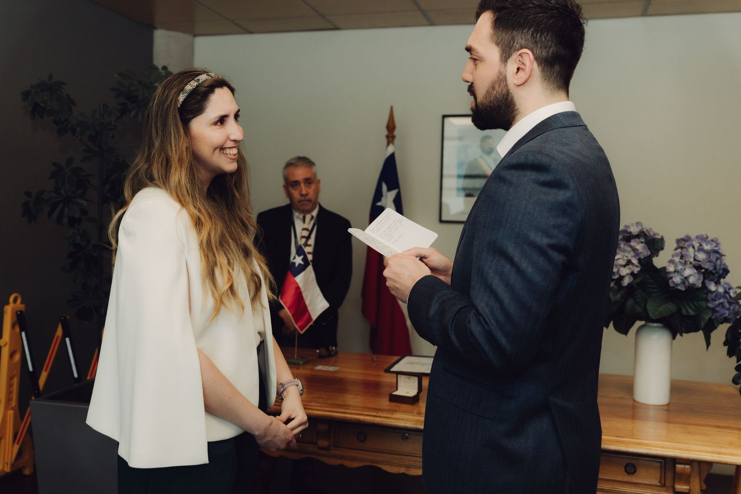 Bearded groom reads wedding vows to smiling bride during civil ceremony with Chilean flag backdrop.