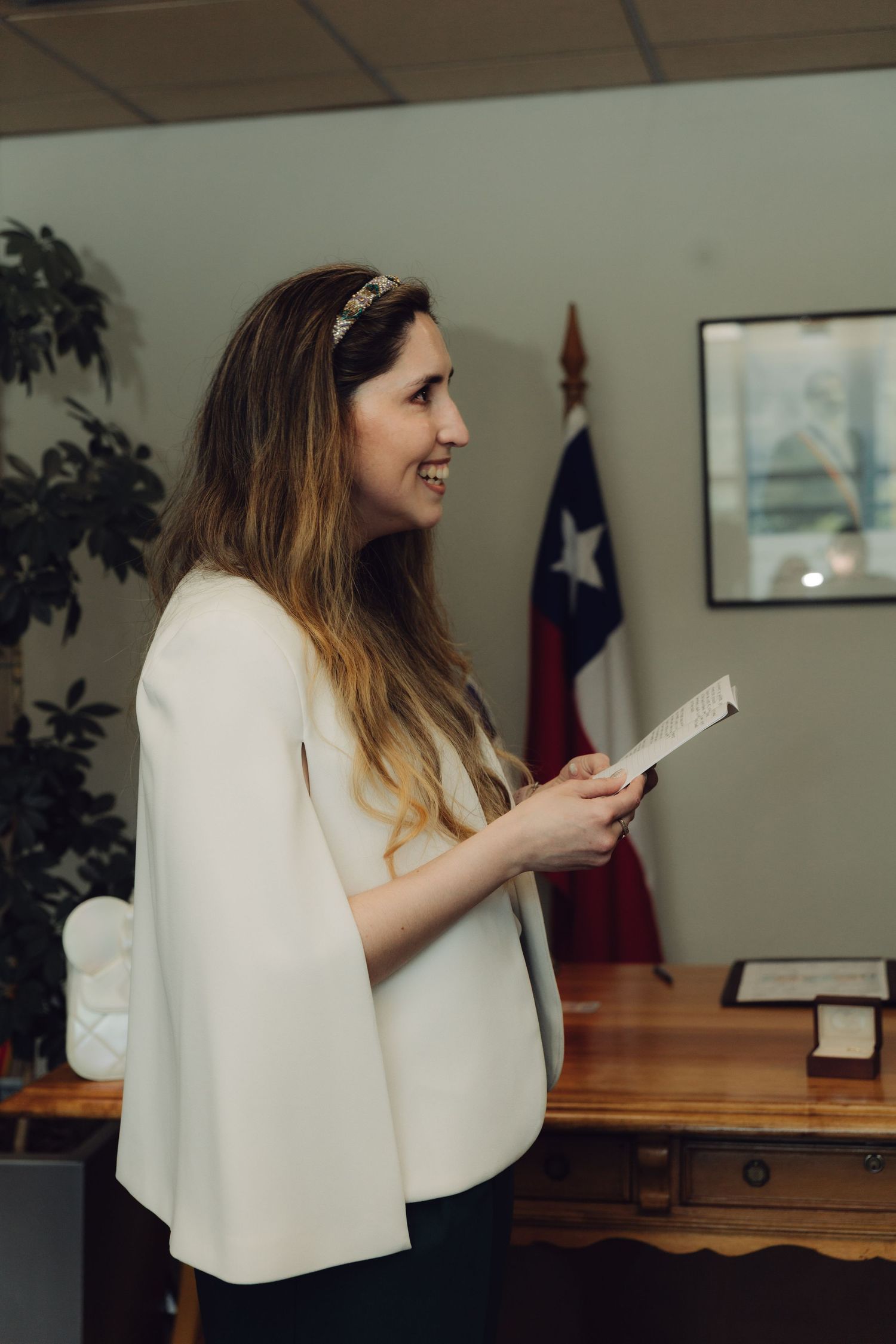 Smiling bride in white cape holds vows paper during civil wedding ceremony with Chilean flag behind her.