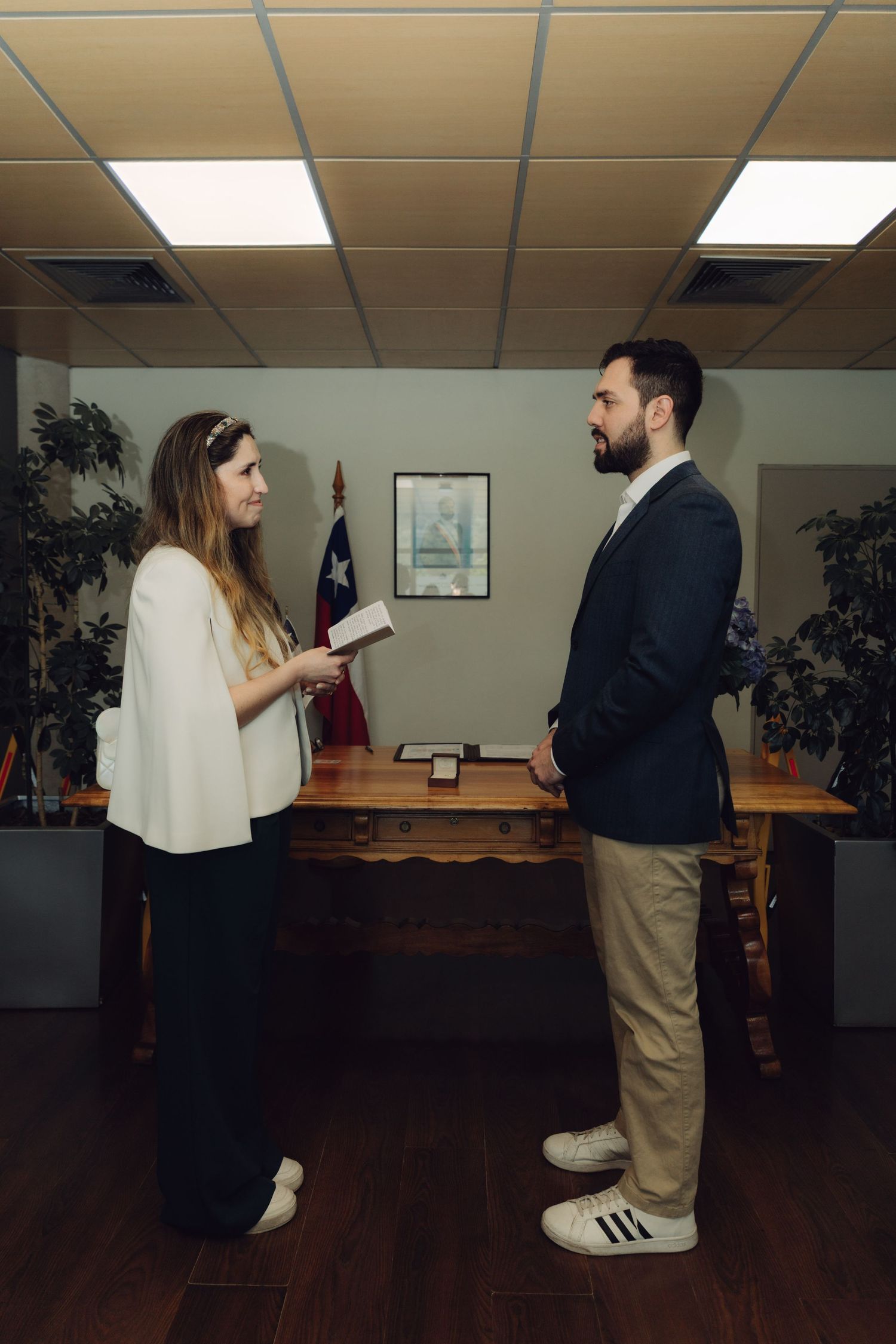 Bride and groom exchange vows facing each other in civil ceremony room with Chilean flag behind them.