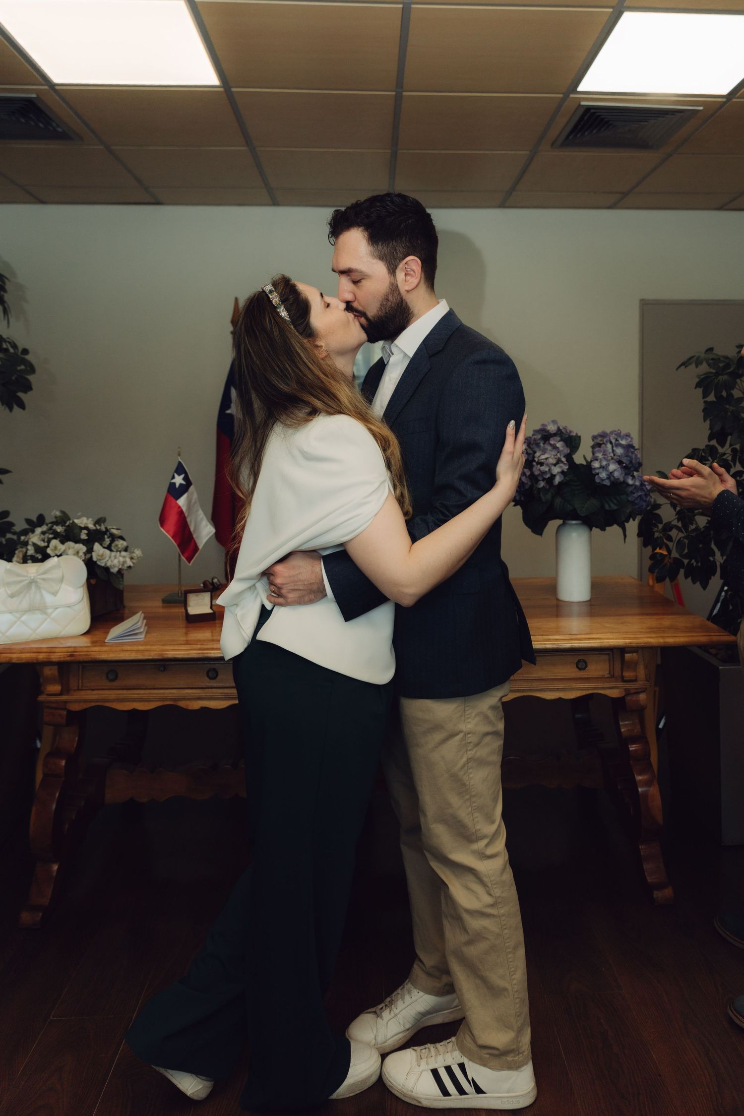 Newlywed couple shares first kiss during civil wedding ceremony with Chilean flag and flowers in background.