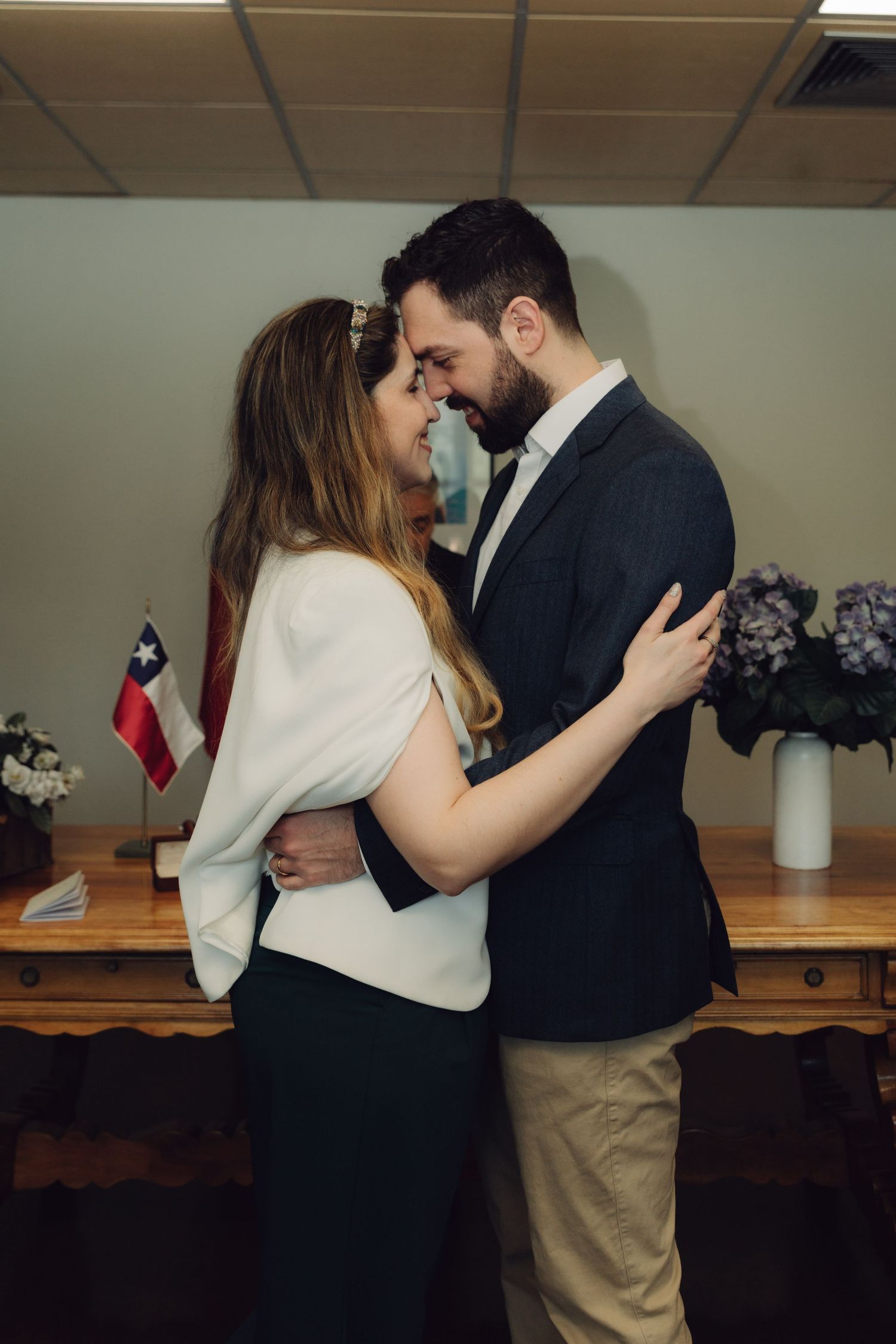Newlywed couple shares intimate embrace after civil wedding ceremony with Chilean flag in background.