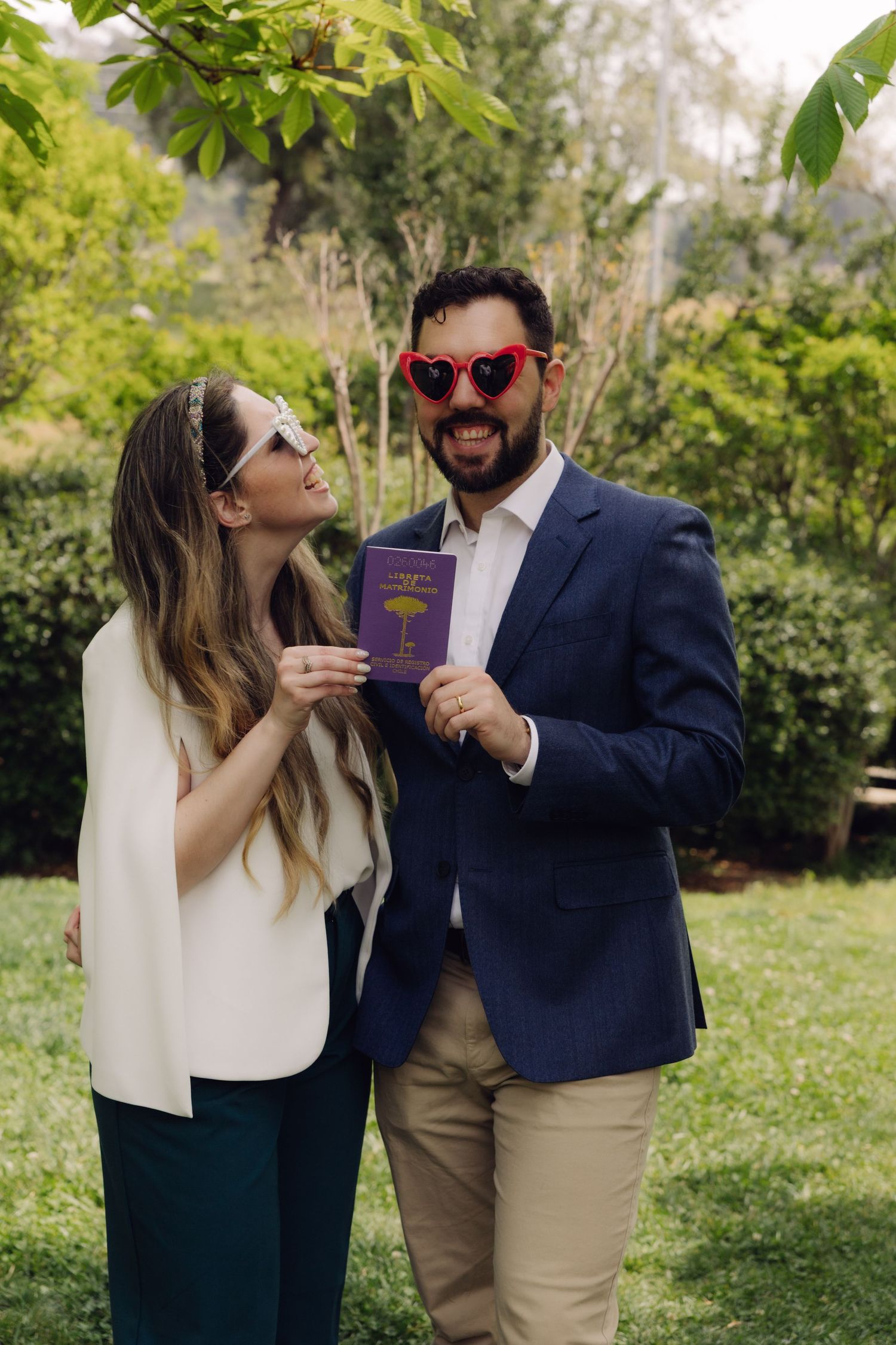 Happy newlywed couple holds marriage booklet outdoors wearing heart sunglasses celebrating after civil wedding.