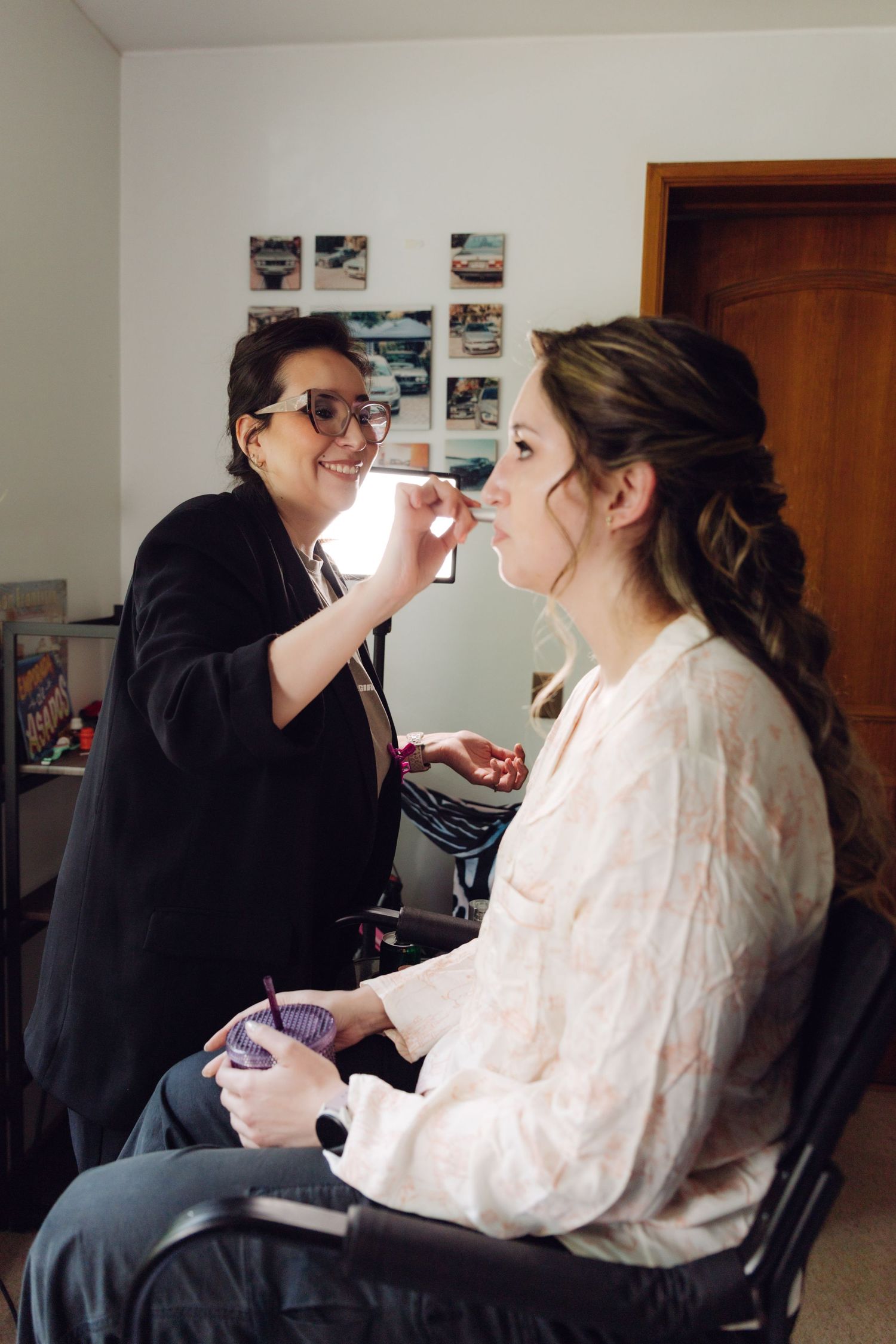 Makeup artist smiling while applying makeup to bride in robe during wedding preparation session.
