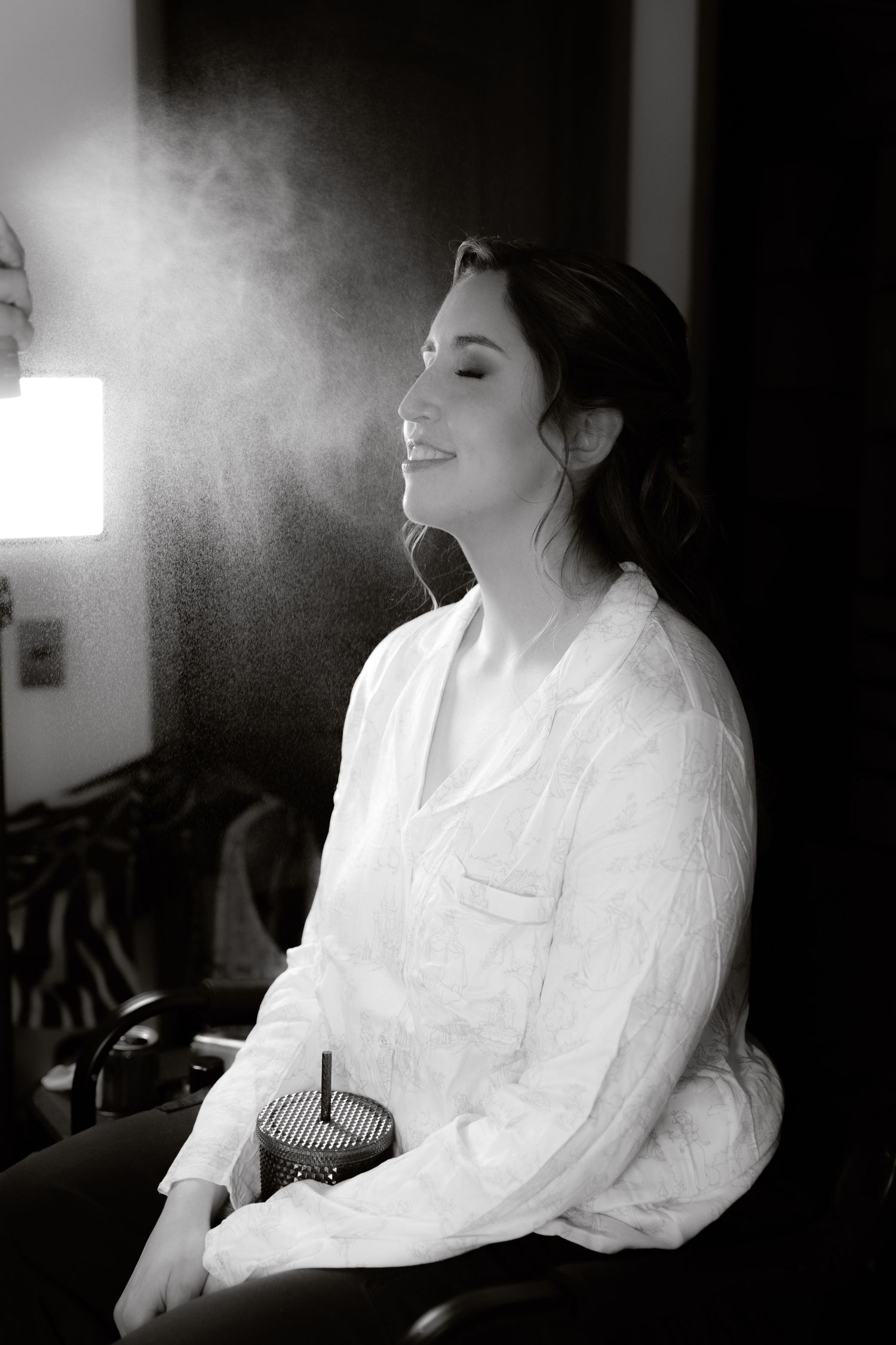 Black and white photo of bride smiling as makeup setting spray is applied.