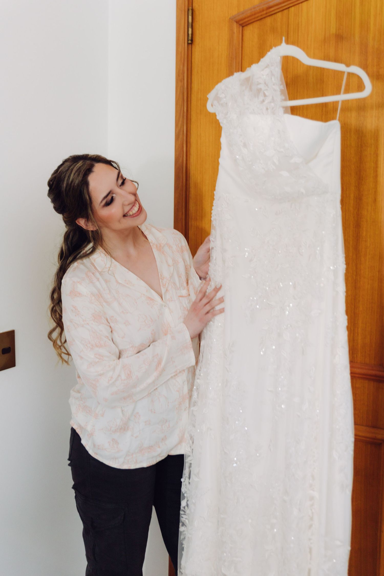 Happy bride in robe admiring her white lace wedding dress on a hanger.