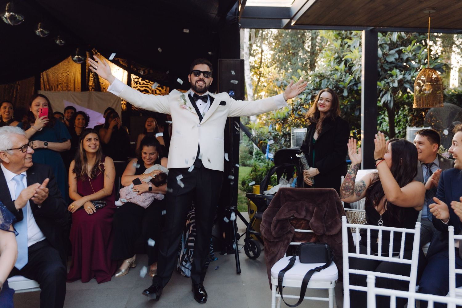 Groom in white tuxedo making grand entrance at wedding reception with guests applauding.