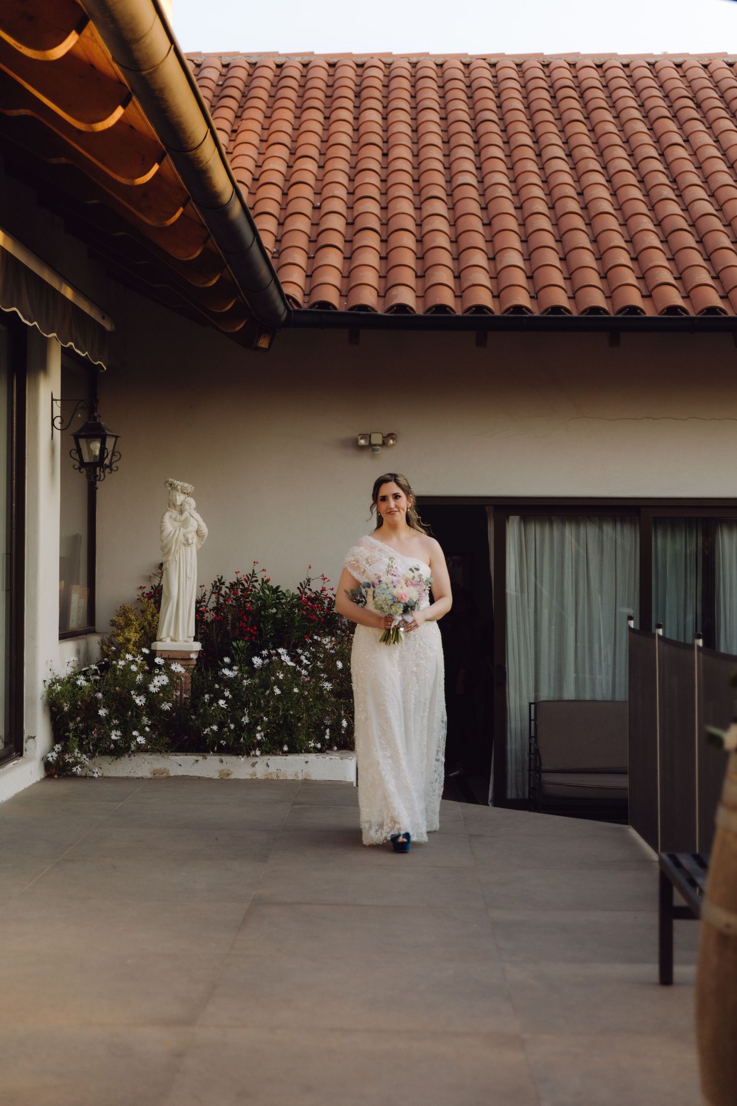 Bride in white lace gown carrying bouquet walking along terrace before ceremony.