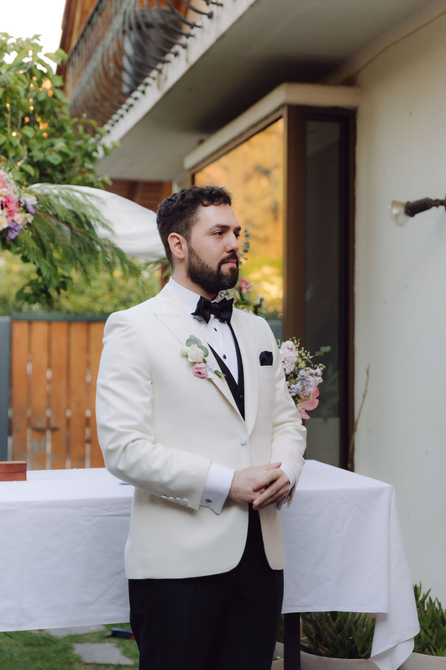 Groom in elegant white tuxedo with floral boutonniere standing at outdoor wedding altar.
