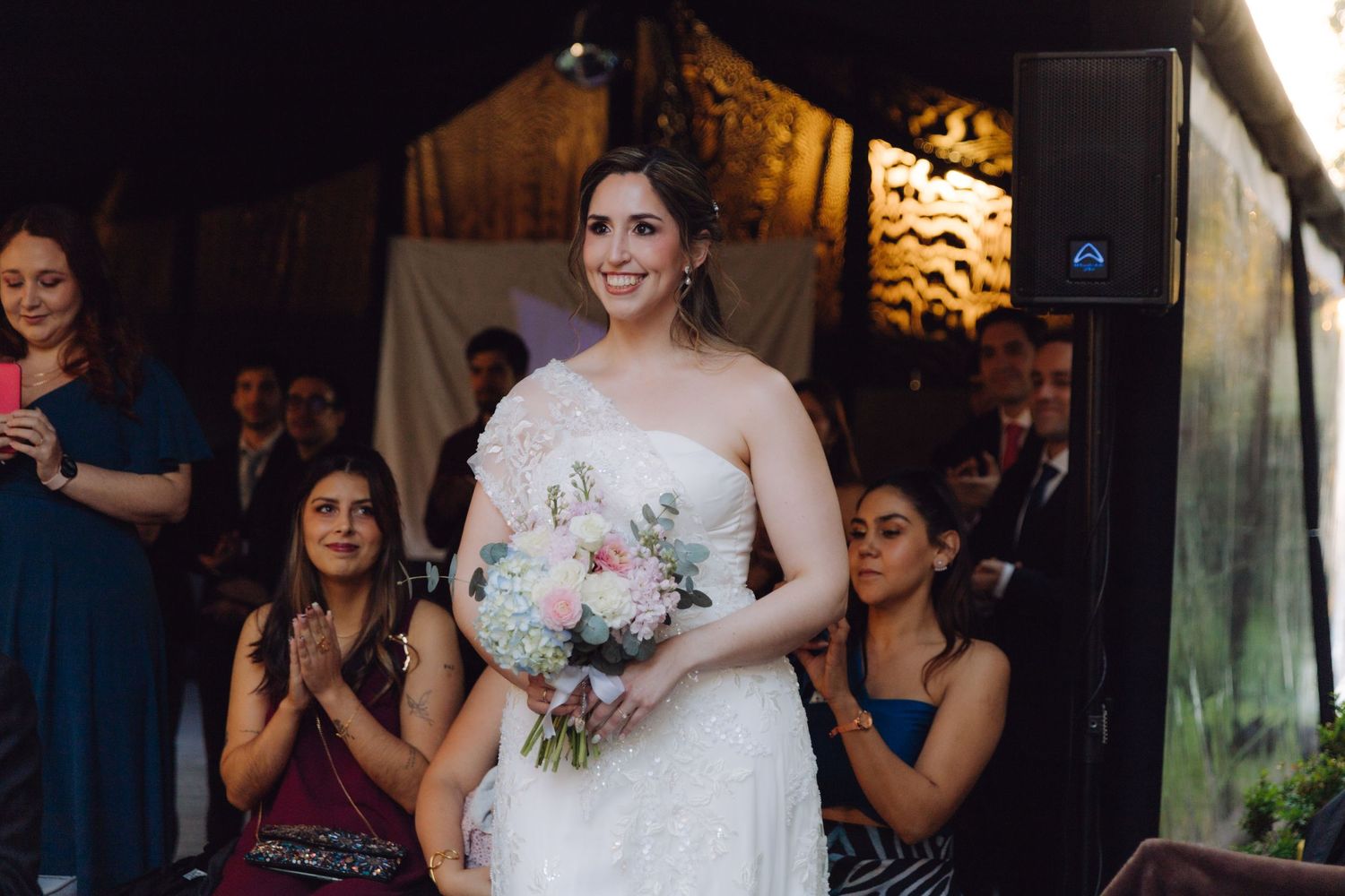 Smiling bride walking down the aisle in white gown holding a pink bouquet.