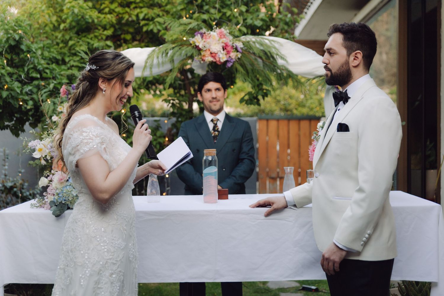 Bride reads her vows into a microphone while groom listens during an outdoor garden wedding ceremony.