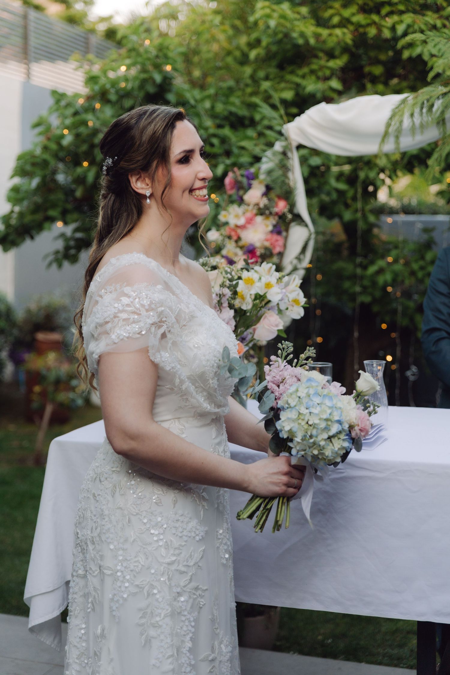 Smiling bride in a lace wedding gown holds a bouquet of pastel flowers during an outdoor ceremony.