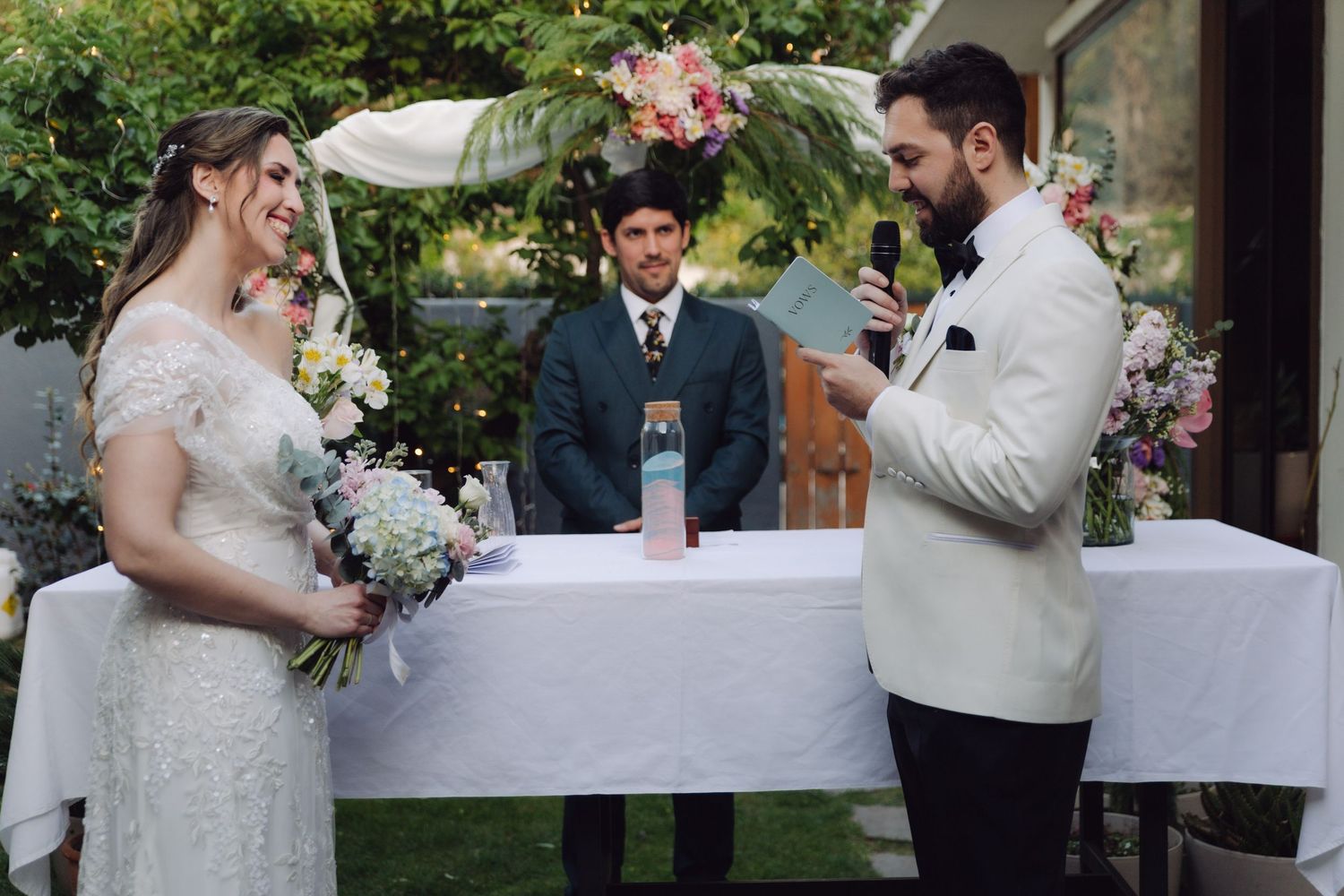 Groom reads his vows to the bride during a lush garden wedding ceremony with floral backdrop.