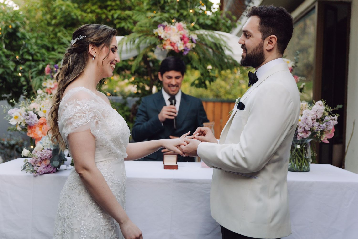 Bride and groom exchange rings while smiling at each other during an outdoor garden wedding ceremony.