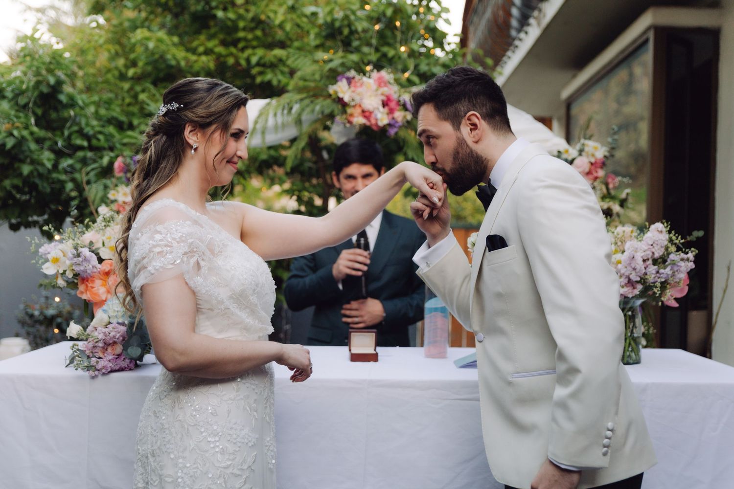 Groom kisses the brides hand during a joyful outdoor wedding ceremony with colorful floral arrangements.