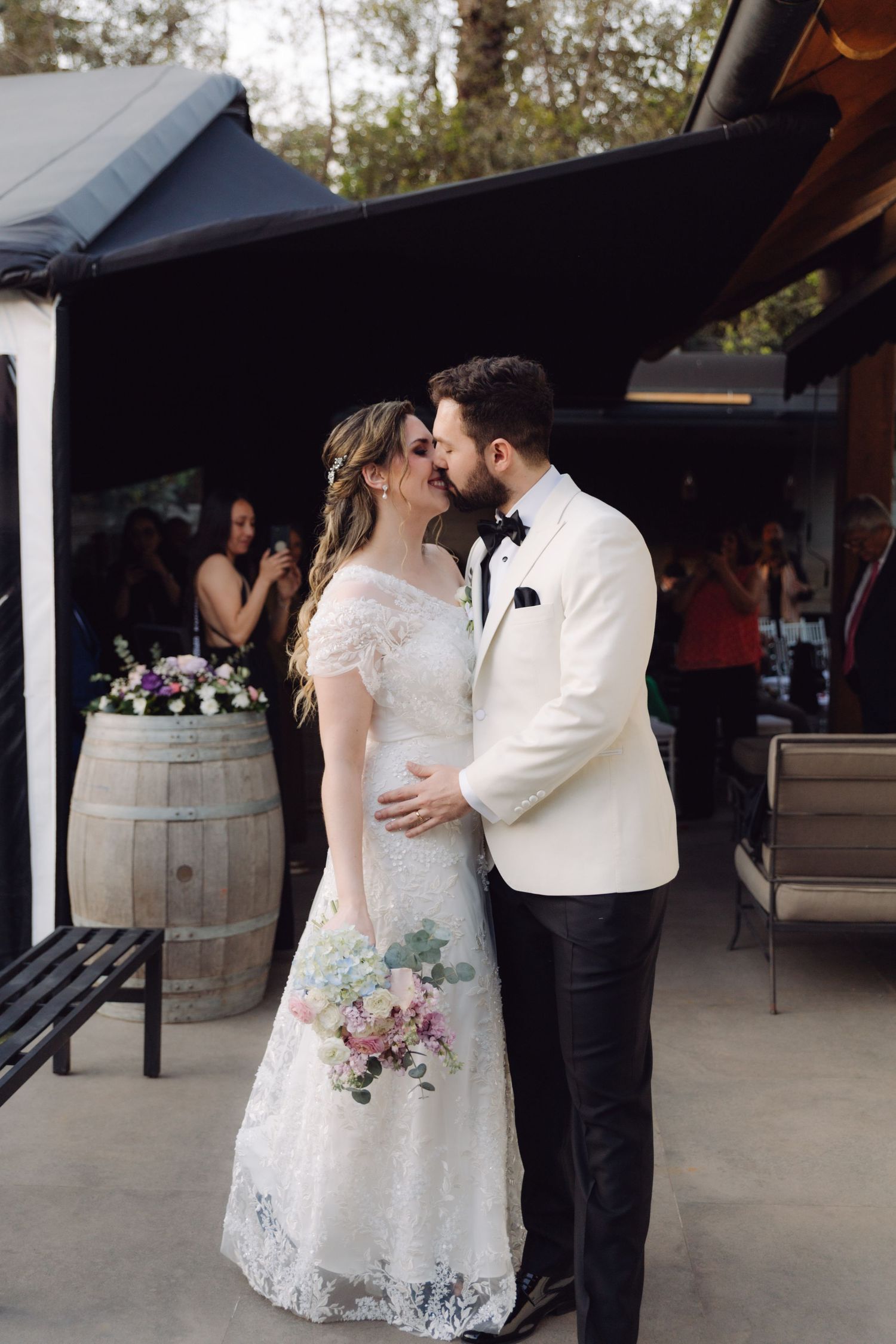 Bride and groom share a romantic kiss at their outdoor wedding reception, she holds a pastel bouquet.