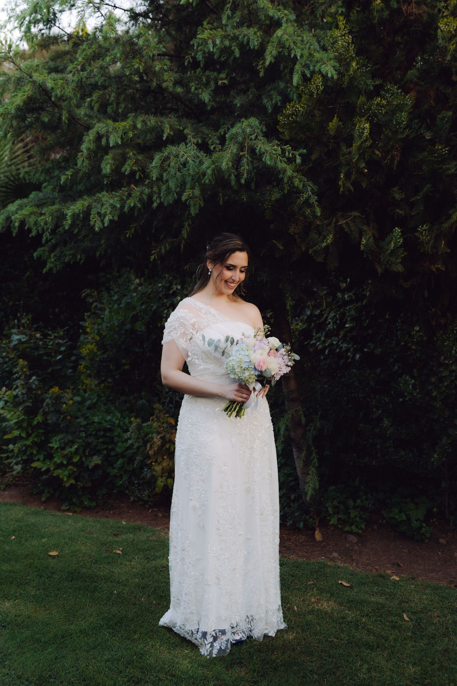 Elegant bride in lace off-shoulder wedding gown holding a pastel bouquet, standing in a lush garden.