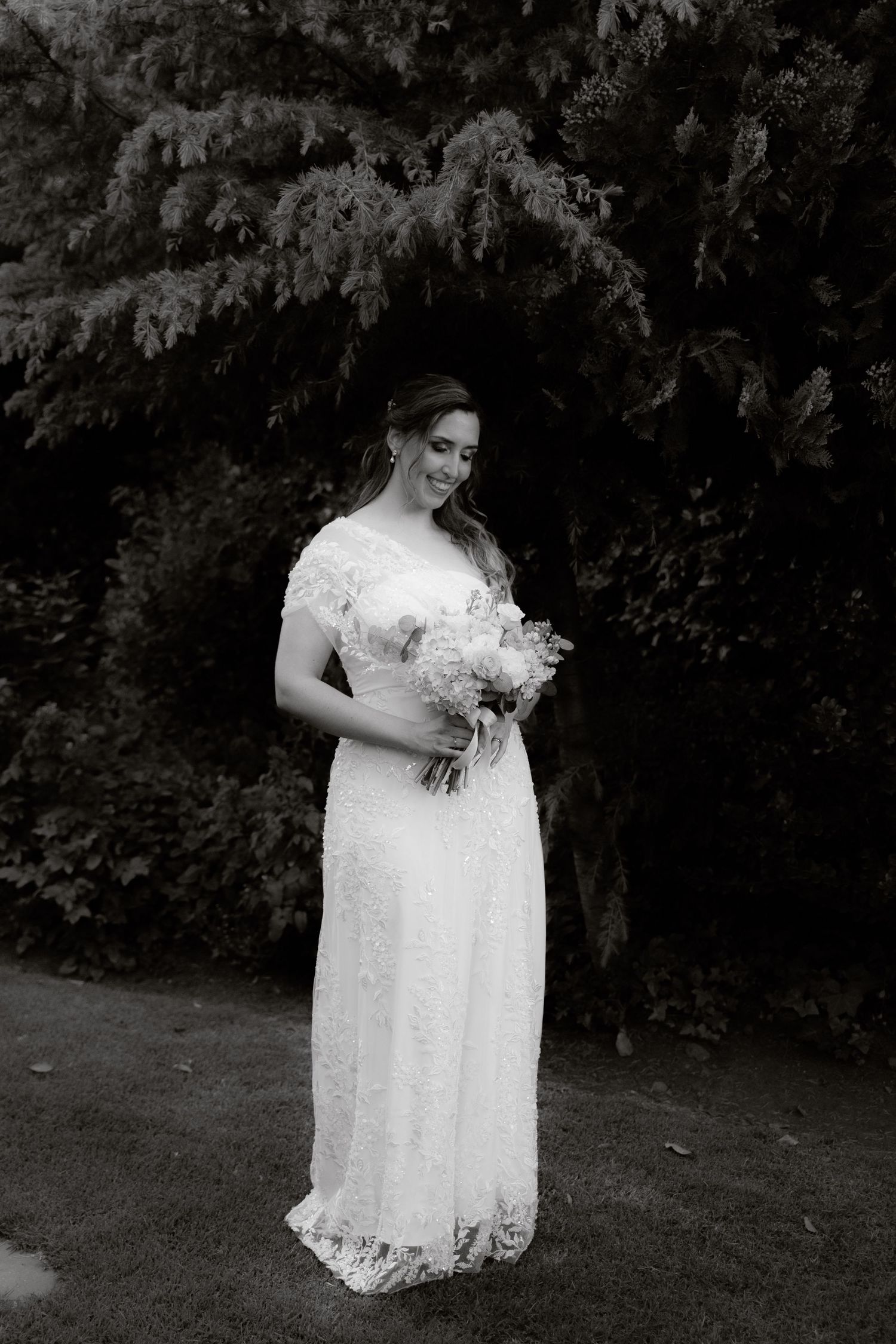 Black and white portrait of a smiling bride in lace gown holding a bouquet outdoors.