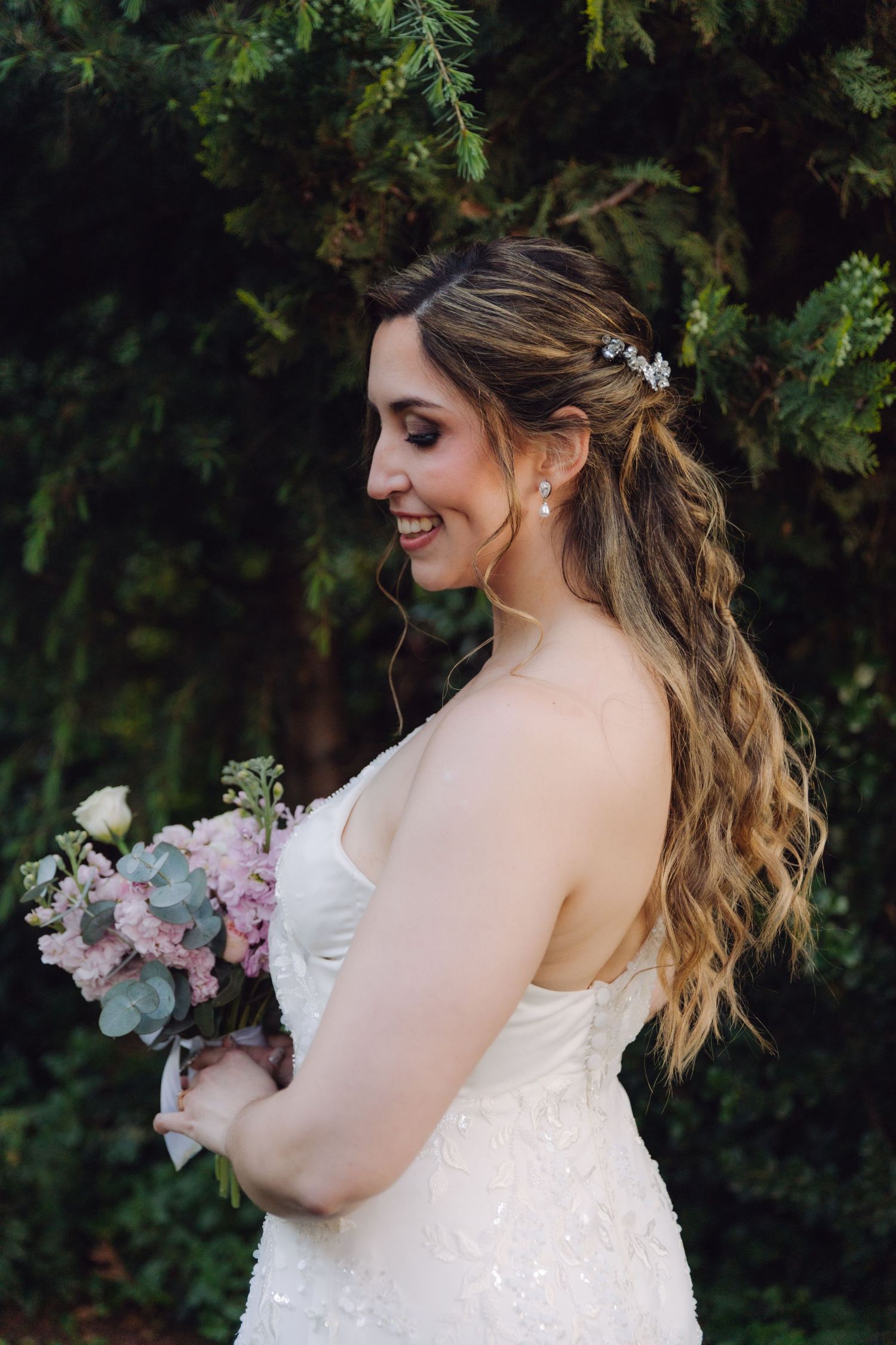 Bride in strapless lace gown smiling with pink floral bouquet in a garden setting.