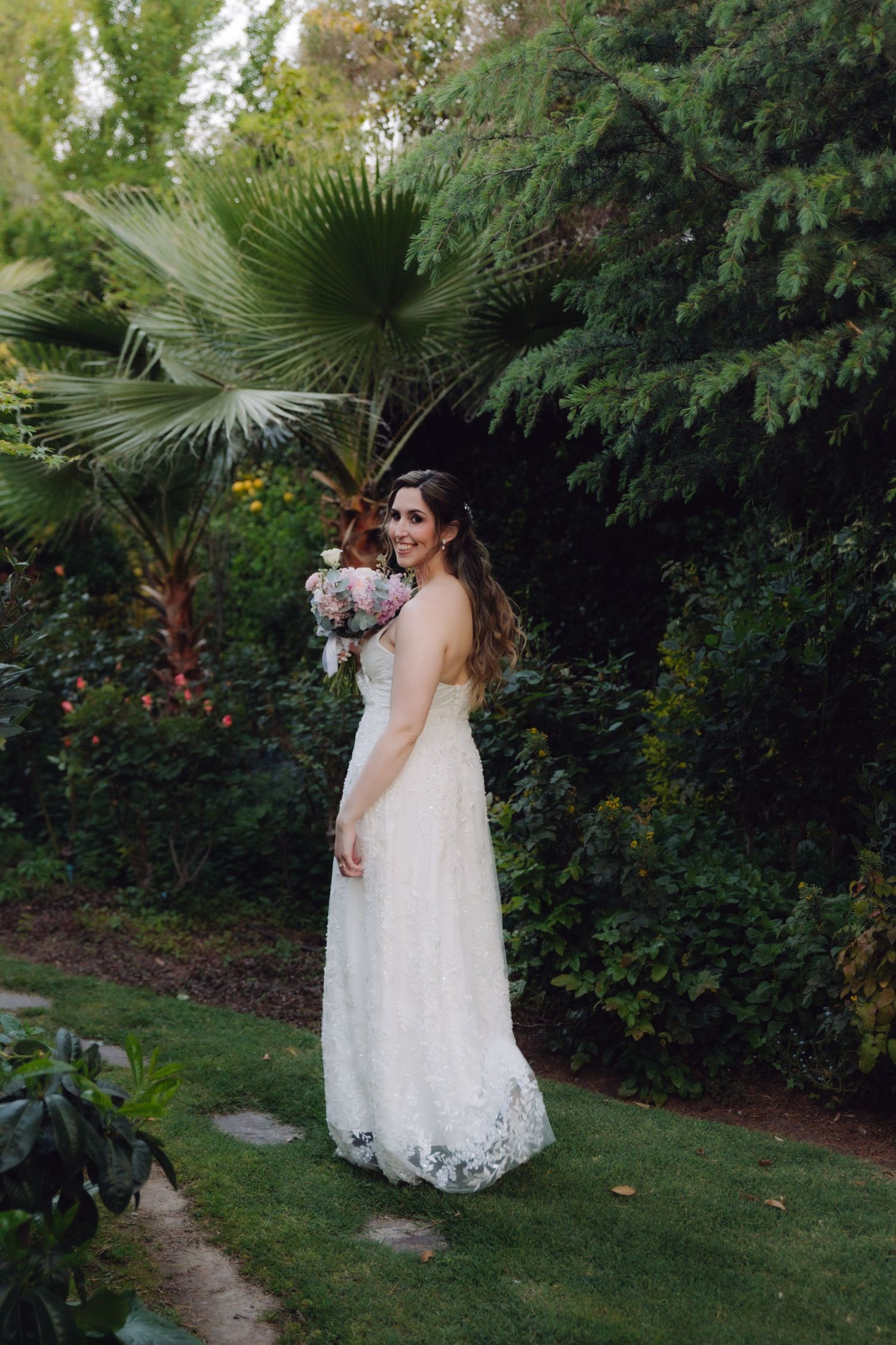 Bride in lace wedding gown holding bouquet while standing on green lawn with tropical plants.