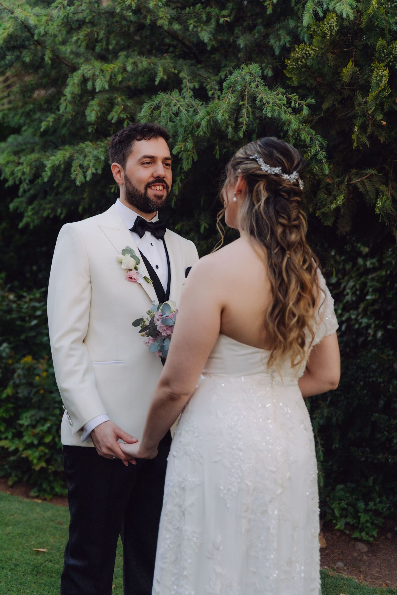 Smiling groom in white tuxedo holding bride's hands during outdoor wedding portrait session.