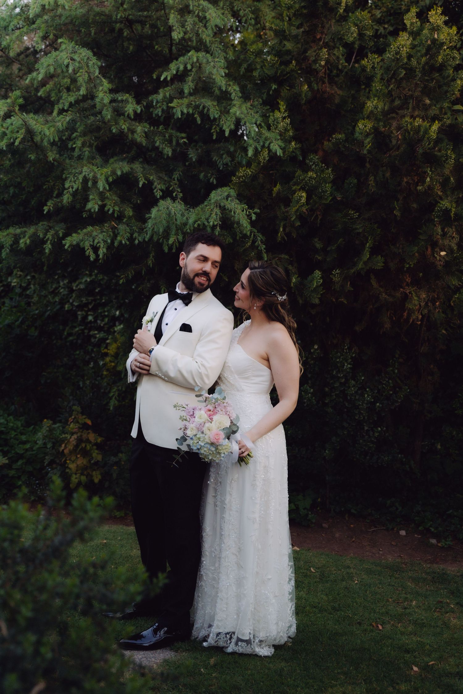 Groom in white tuxedo jacket and bride holding pastel bouquet pose together in garden.