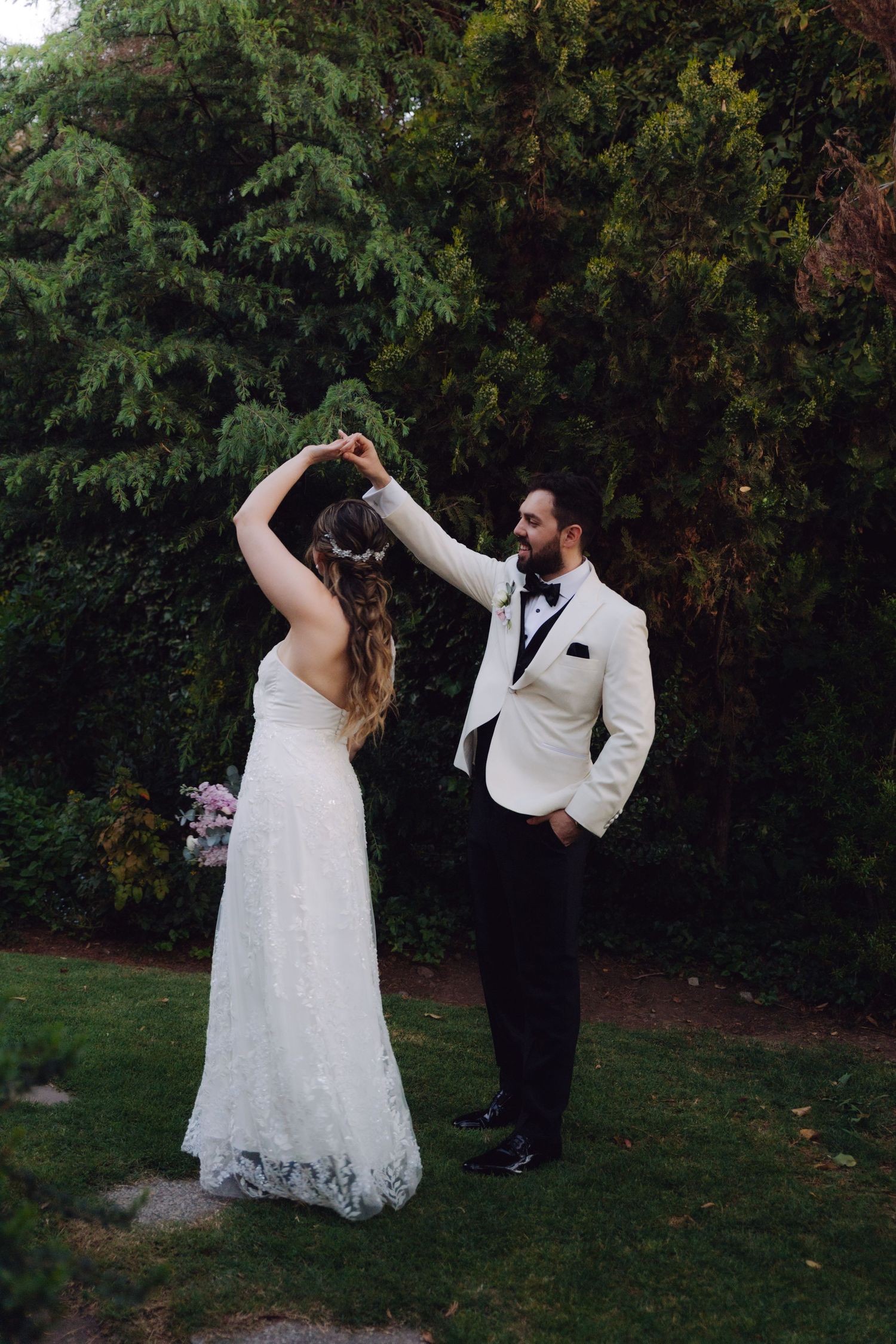 Groom in white tuxedo spins bride in lace wedding dress during outdoor garden photo session.