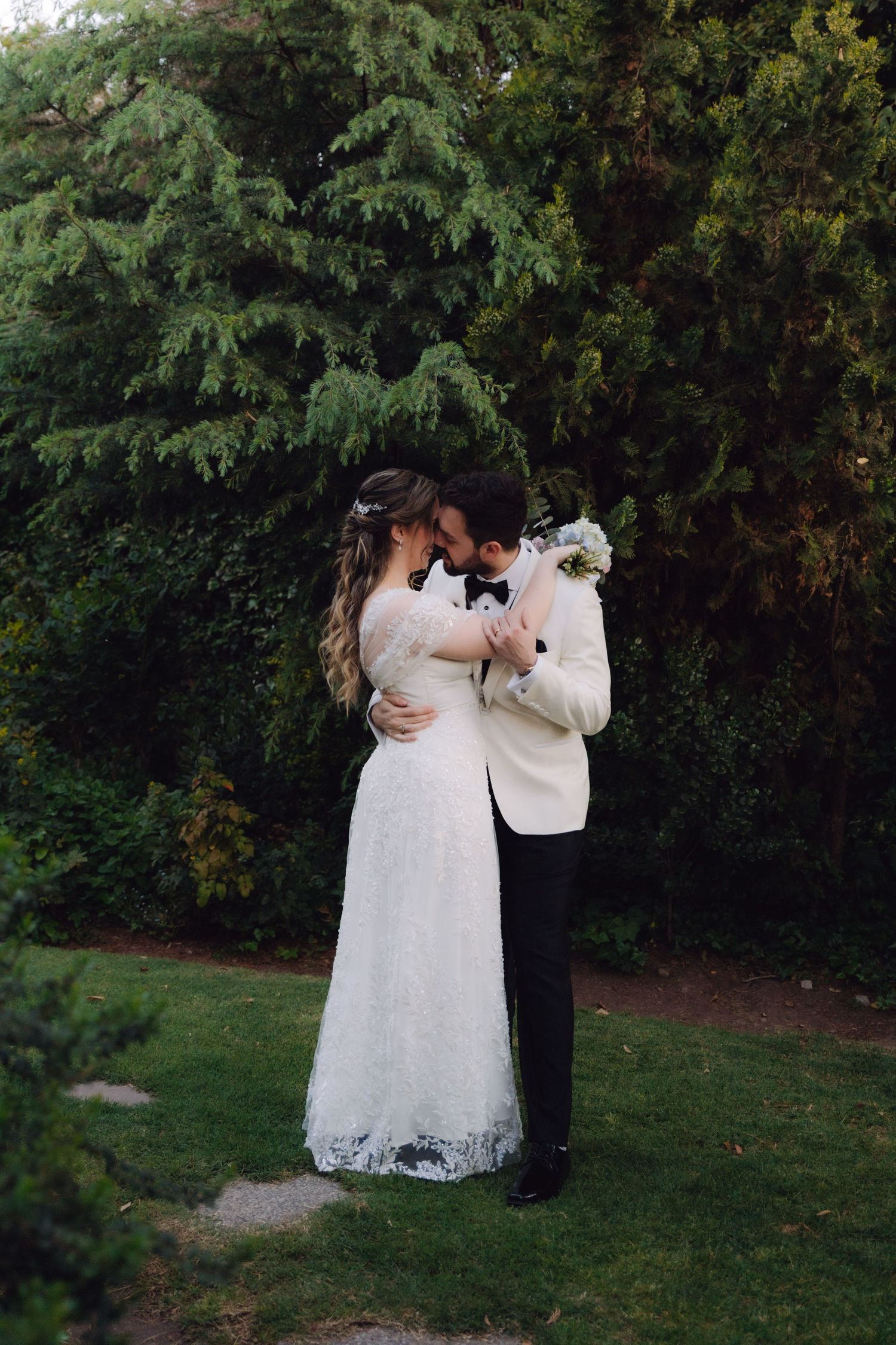 Bride and groom share a close romantic embrace holding bouquet in a lush green garden.