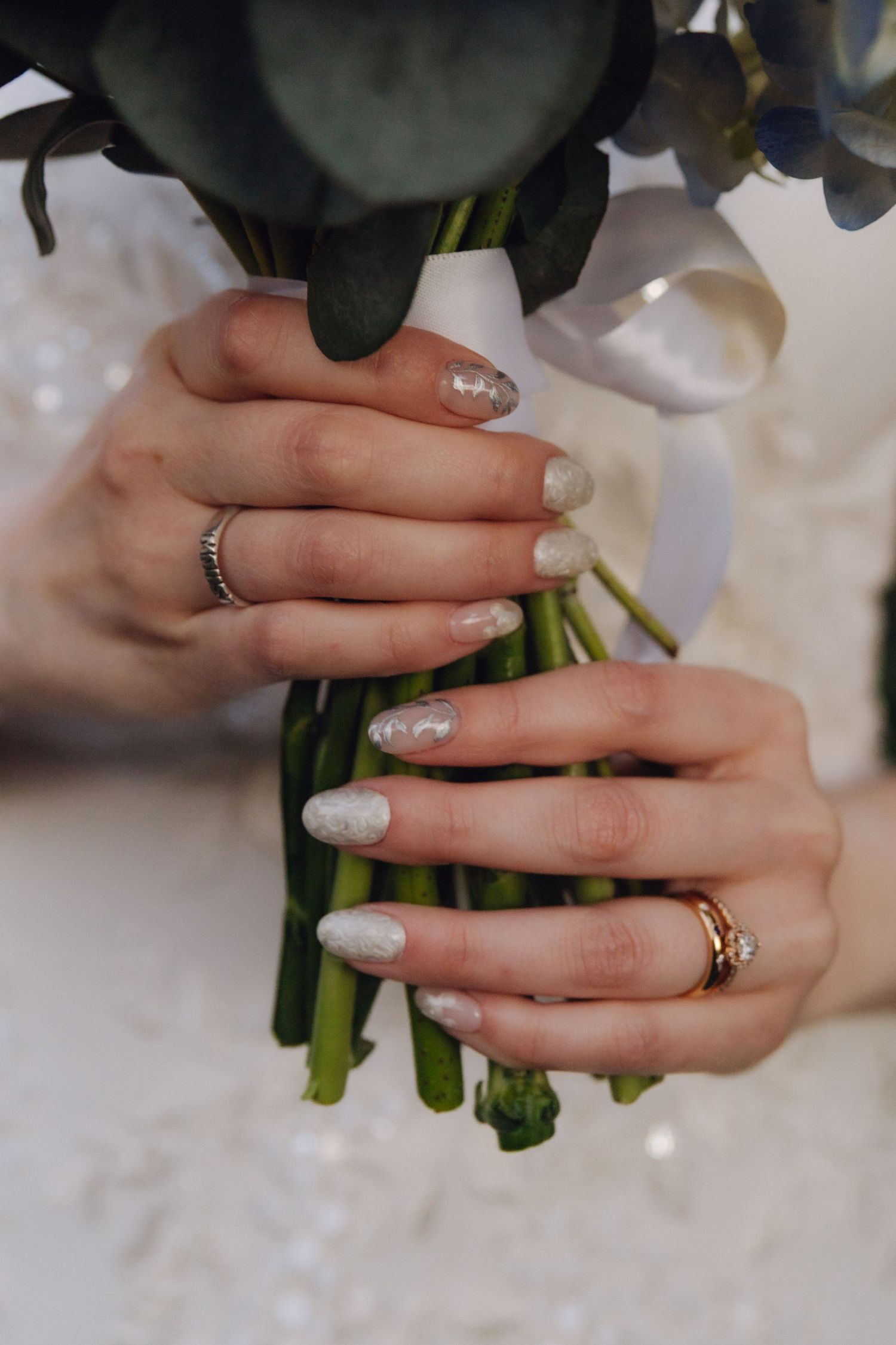 Close-up of bride's hands with pearl manicure and gold rings holding an elegant floral bouquet.