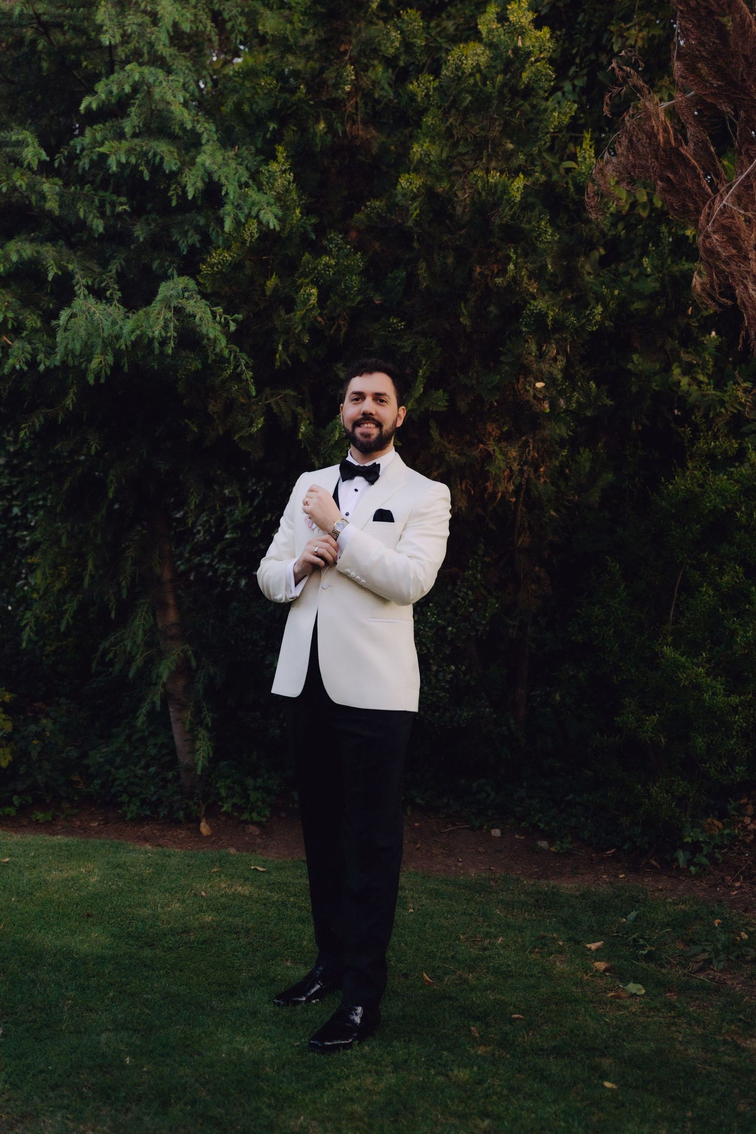 Confident groom in white tuxedo jacket and black bow tie poses alone in lush garden.