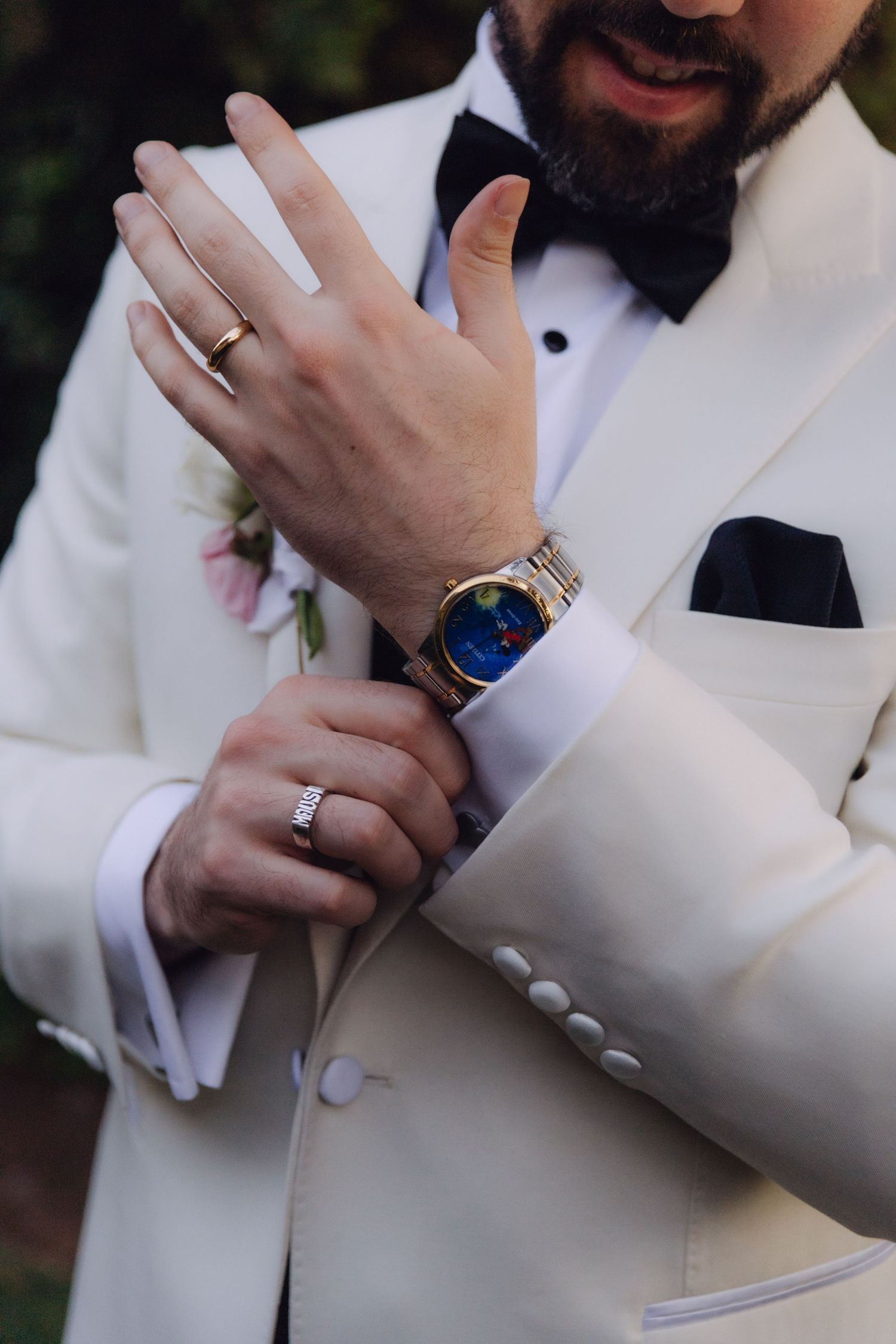 Groom adjusting gold watch with colorful dial, wearing wedding ring and white tuxedo with black bow tie.