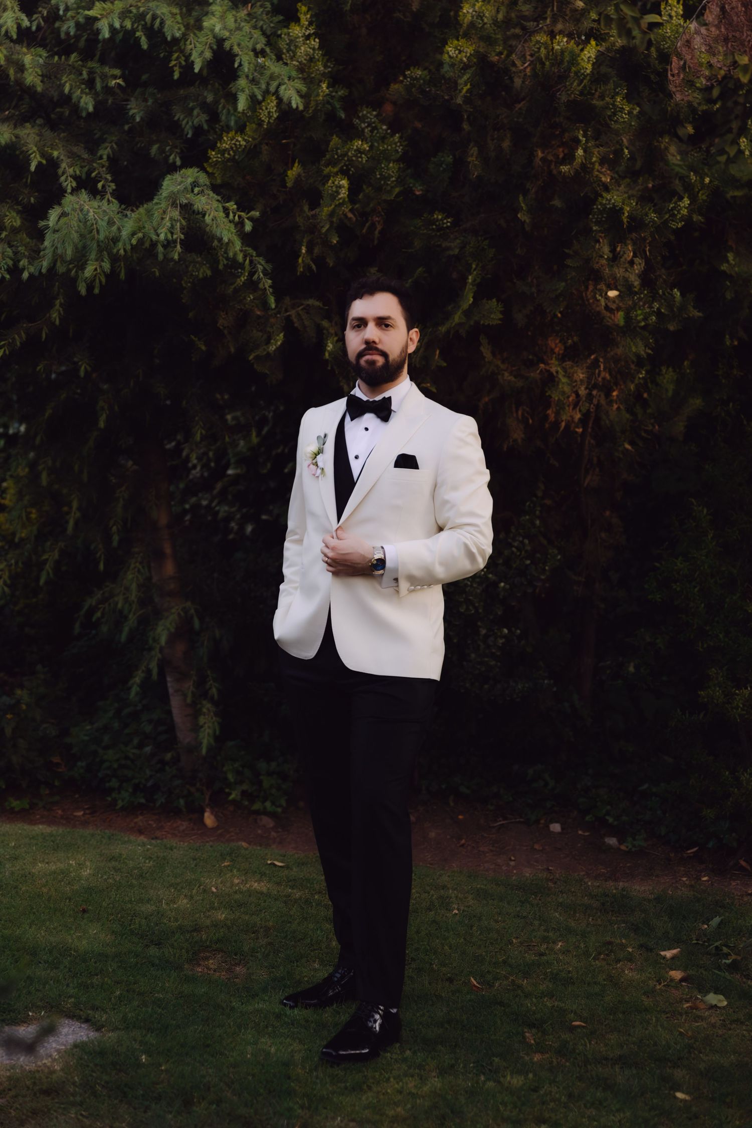 Elegant groom in white tuxedo jacket with black bow tie and pocket square standing in garden.