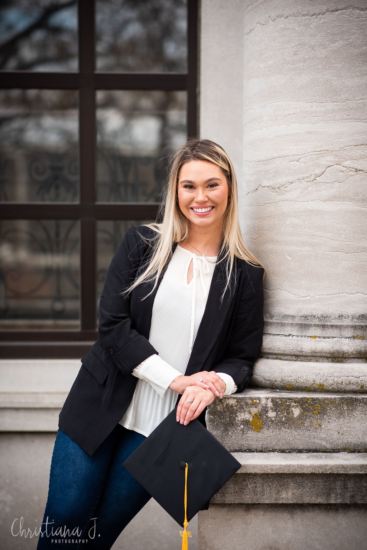 Blonde woman in black blazer holding graduation cap beside a large stone column.