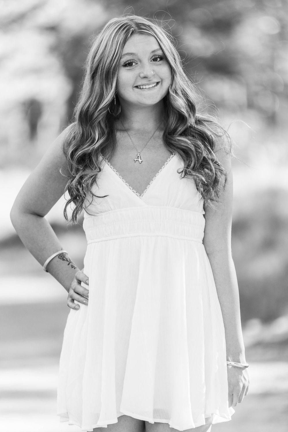 Smiling young woman with long wavy hair wearing a white sundress and necklace, posing outdoors in black and white photo.