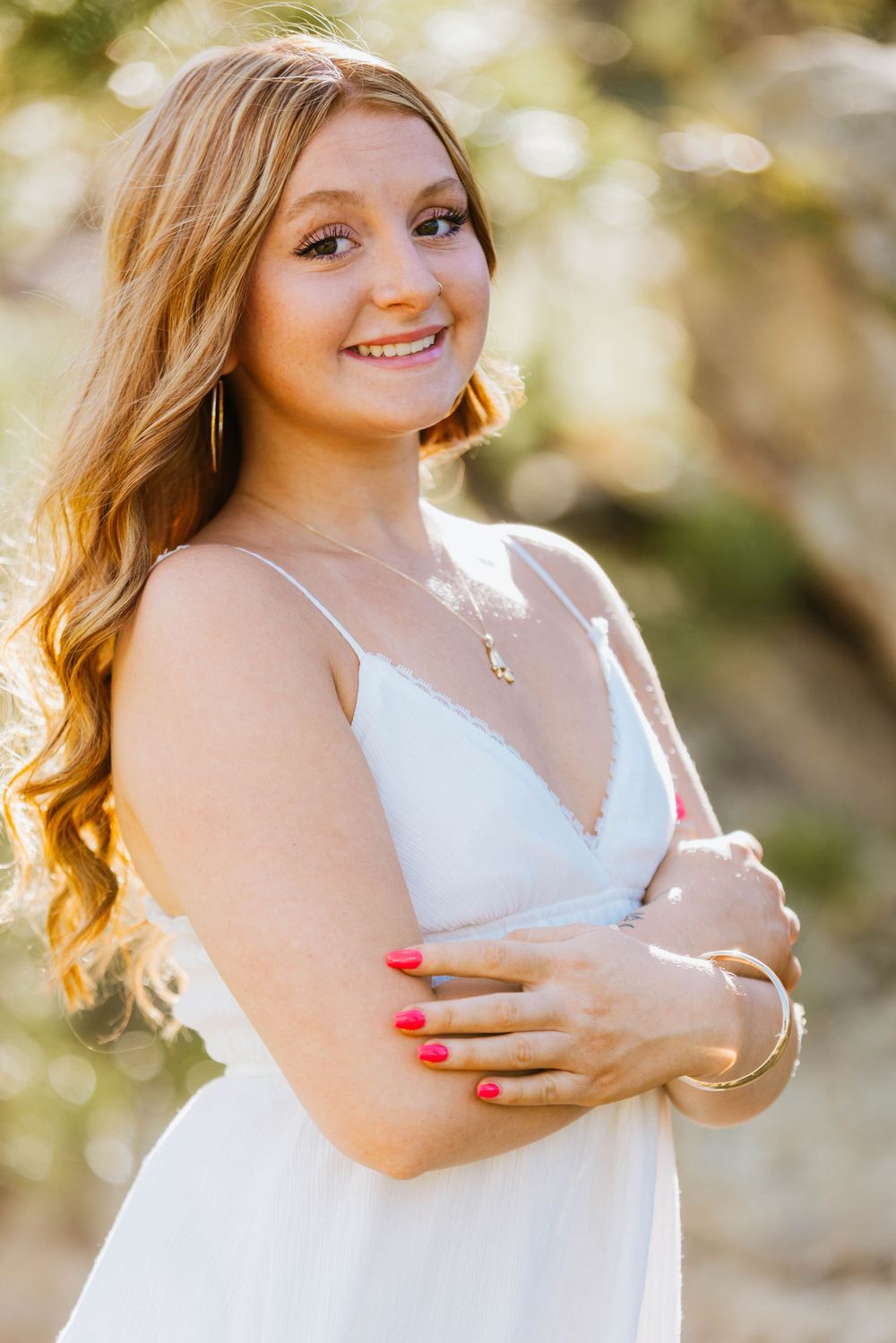 Smiling young woman with long blonde hair wearing a white sundress with arms crossed outdoors.