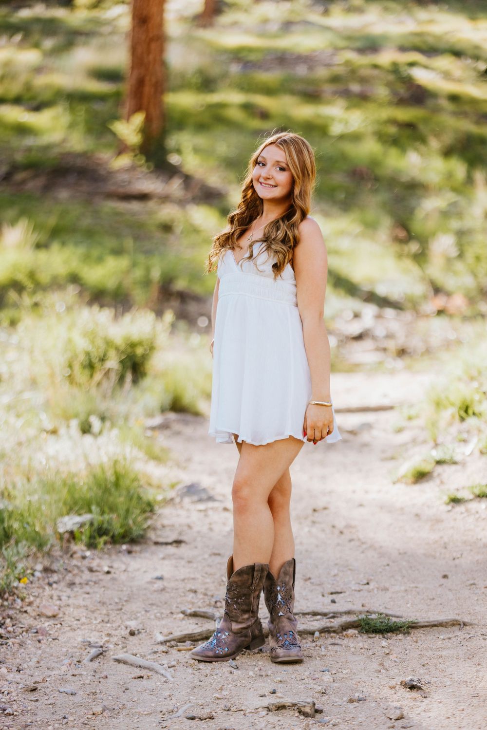 Young woman in white sundress and cowboy boots smiling outdoors on a dirt path surrounded by green trees.