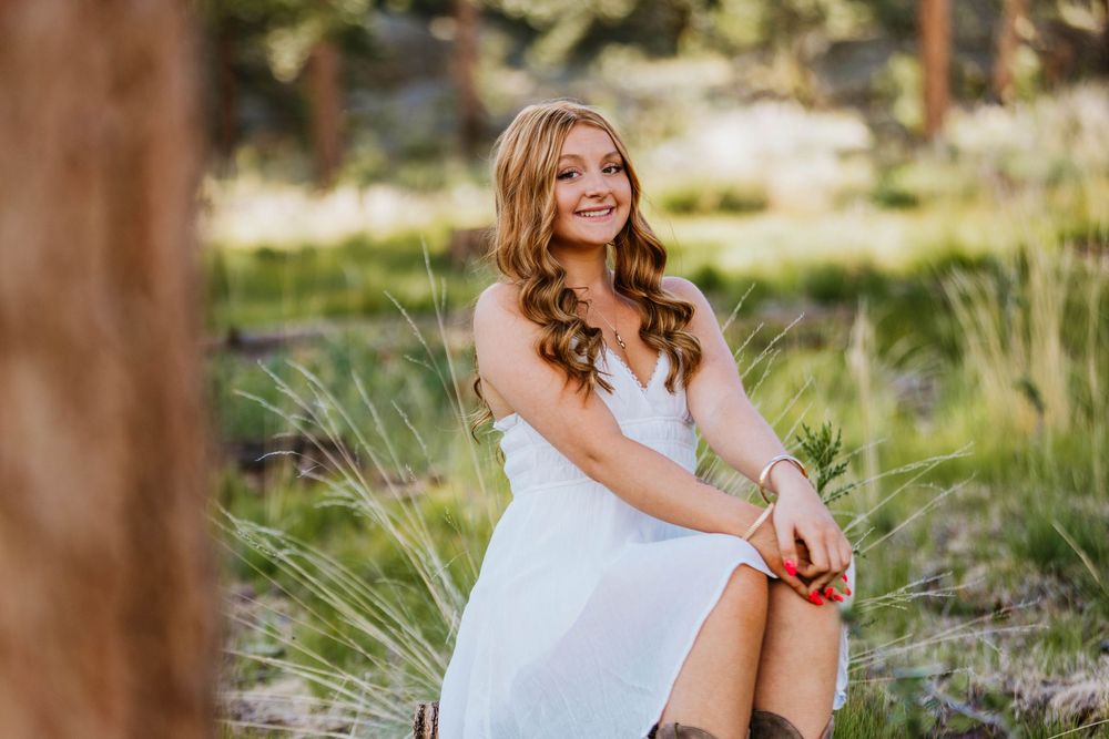 Young woman in white dress sitting outdoors in a forest setting, smiling warmly during a senior portrait session.