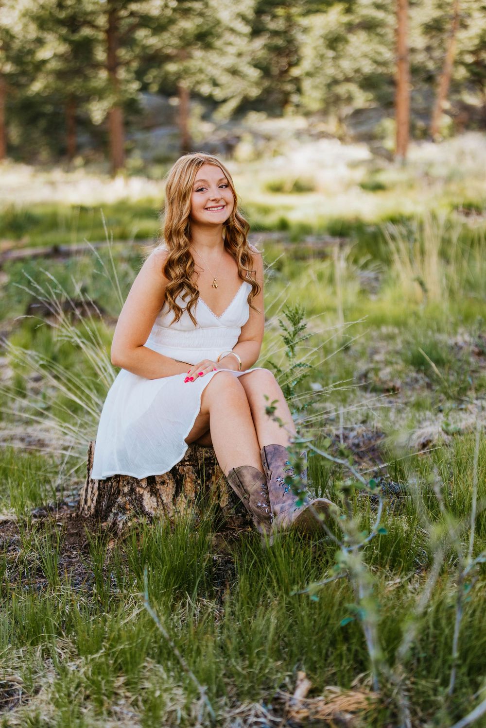Young woman in white dress and cowboy boots sitting on a tree stump in a lush green forest, smiling.